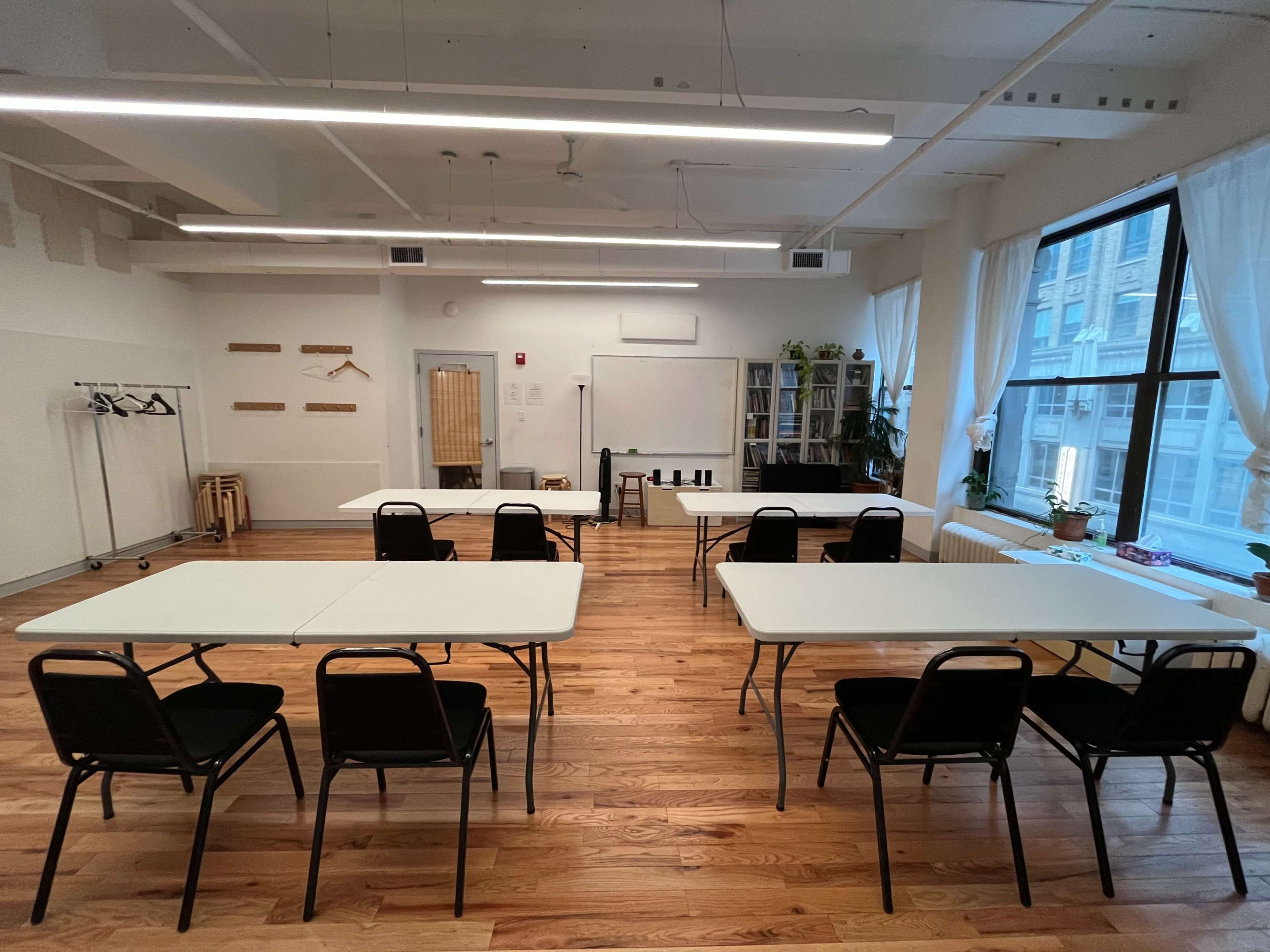 A spacious room with several tables and chairs arranged for a meeting, featuring large windows and wooden flooring.