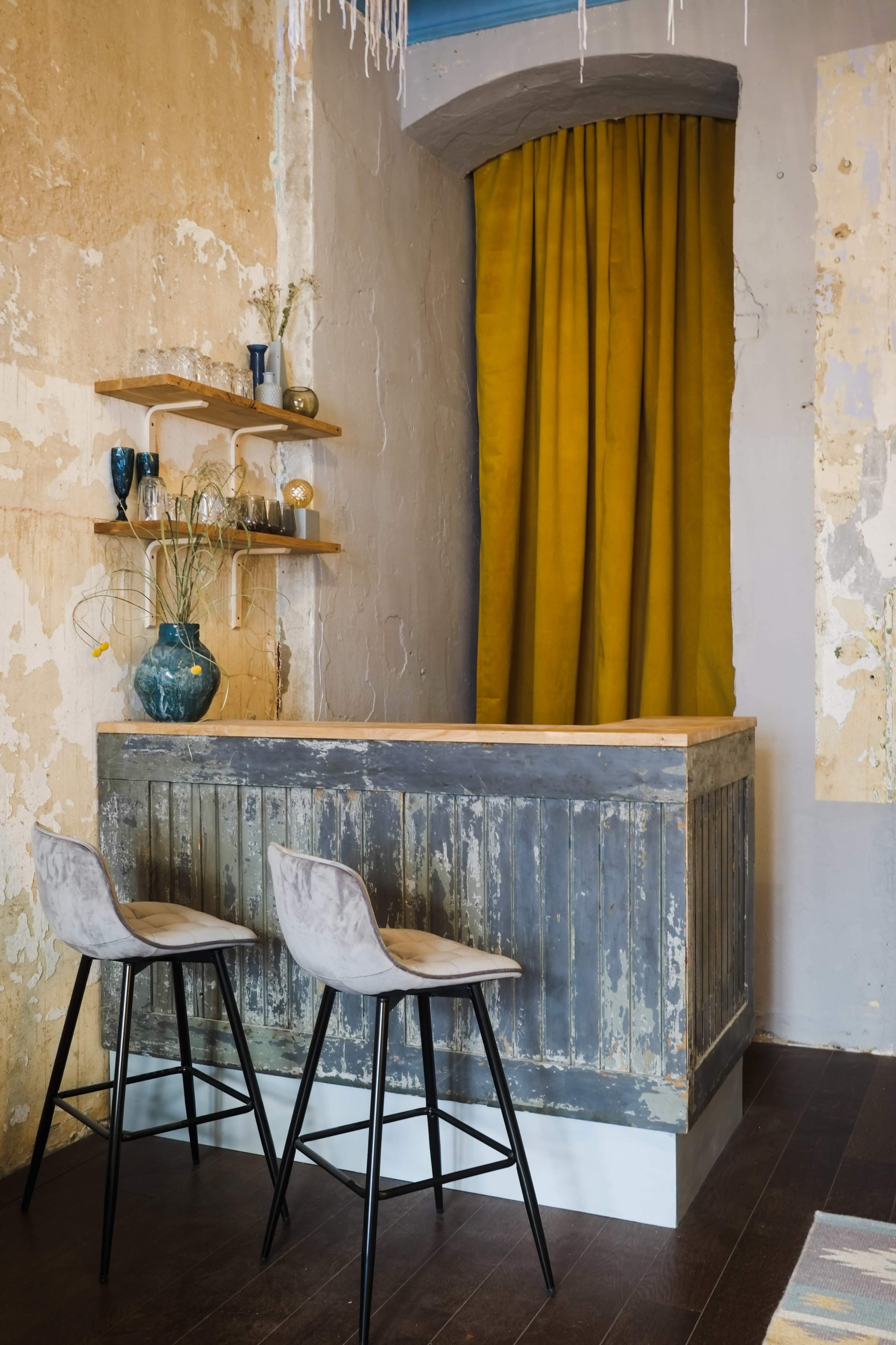 A small bar area with two stools, a wooden counter, and a yellow curtain in a room with textured walls.