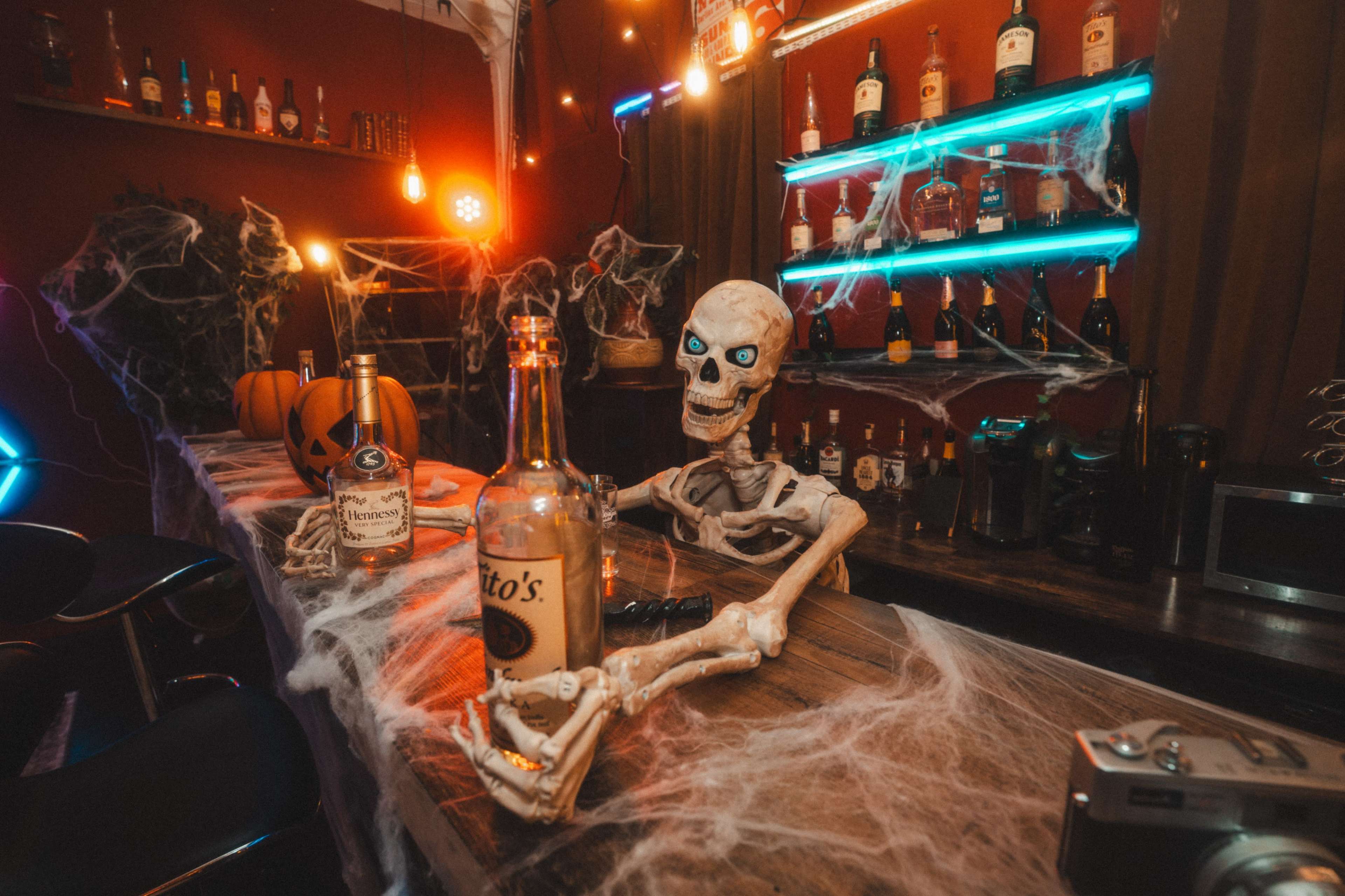 A skeleton sits at a bar adorned with Halloween decorations, including cobwebs and a pumpkin, while various bottles line the shelves in the background.