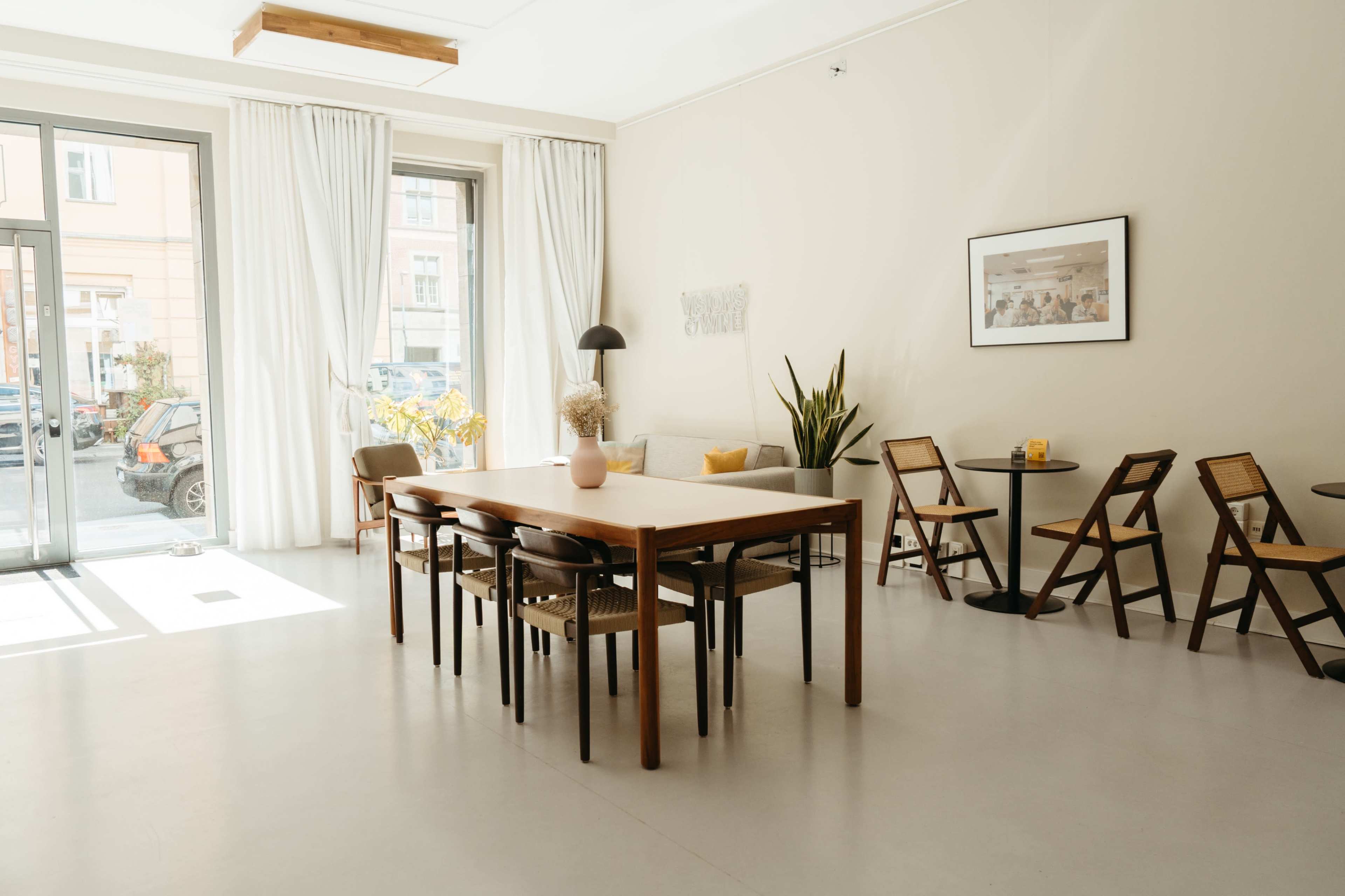 A spacious room features a large wooden dining table surrounded by chairs, with floor-to-ceiling windows letting in natural light.