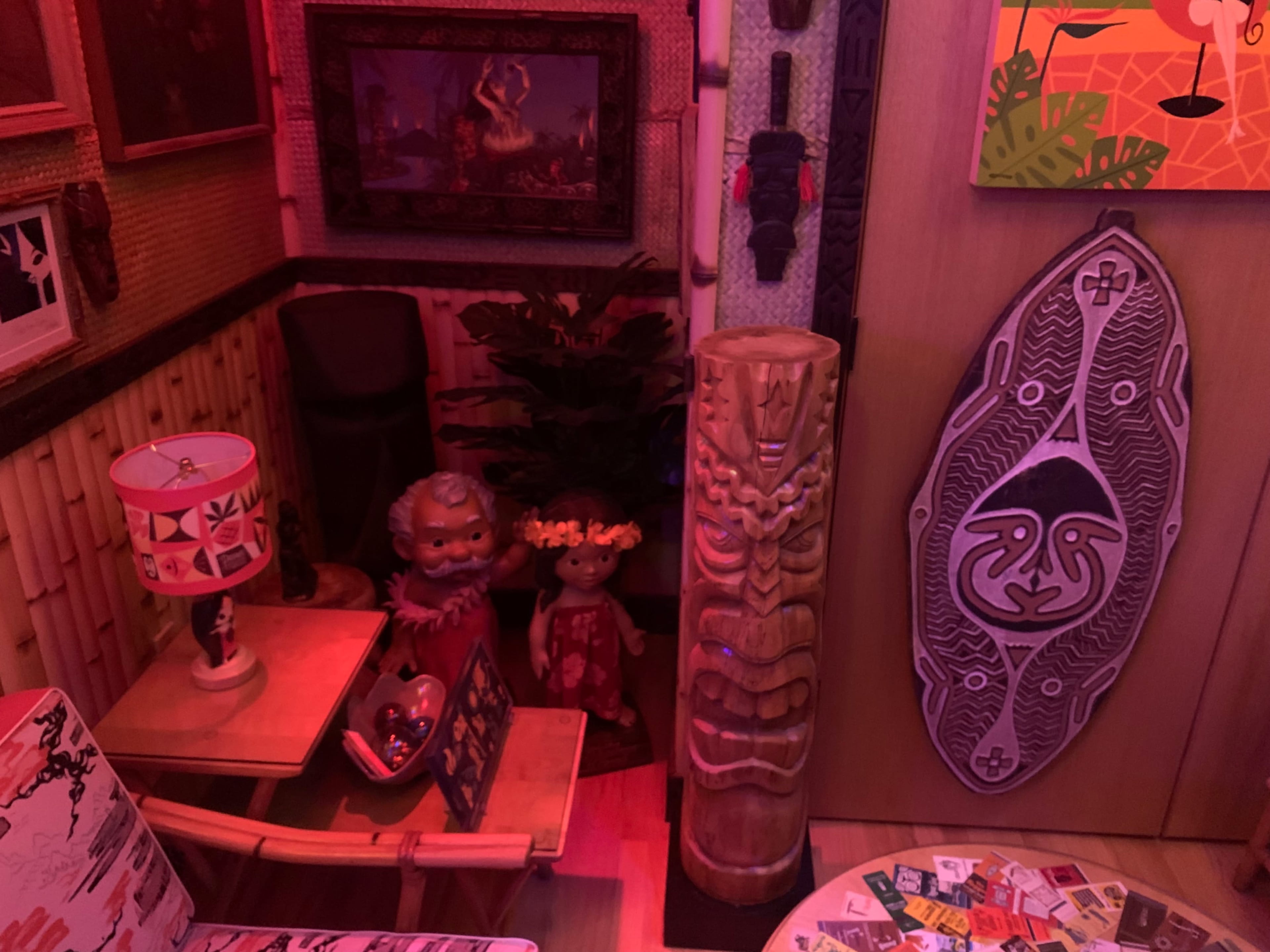The scene features a cozy room adorned with tiki-inspired decor, including carved wooden masks, figurines, and colorful artwork illuminated by red lighting.