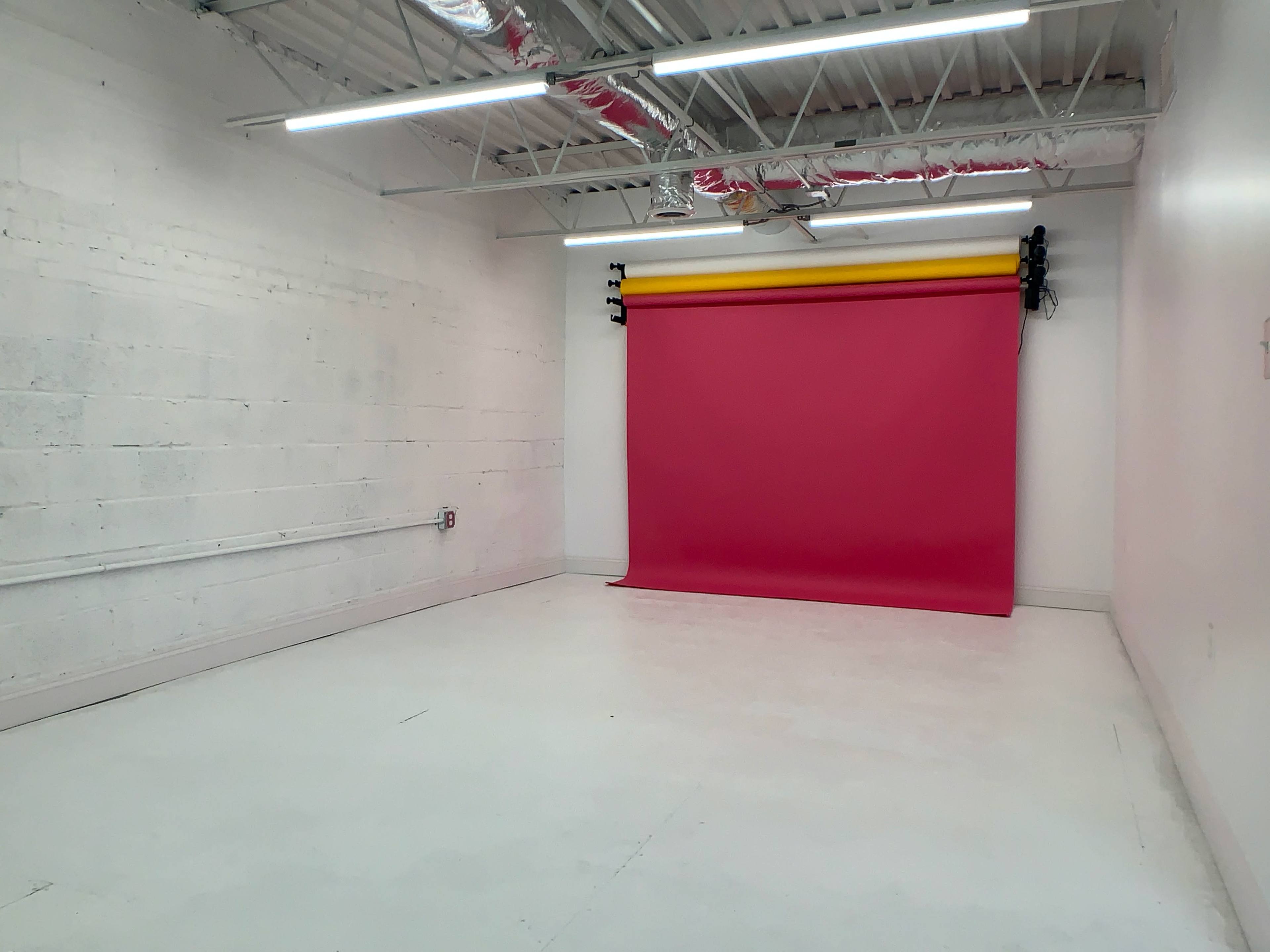 A mostly empty studio space with a pink backdrop and yellow roll at one end against a white wall.