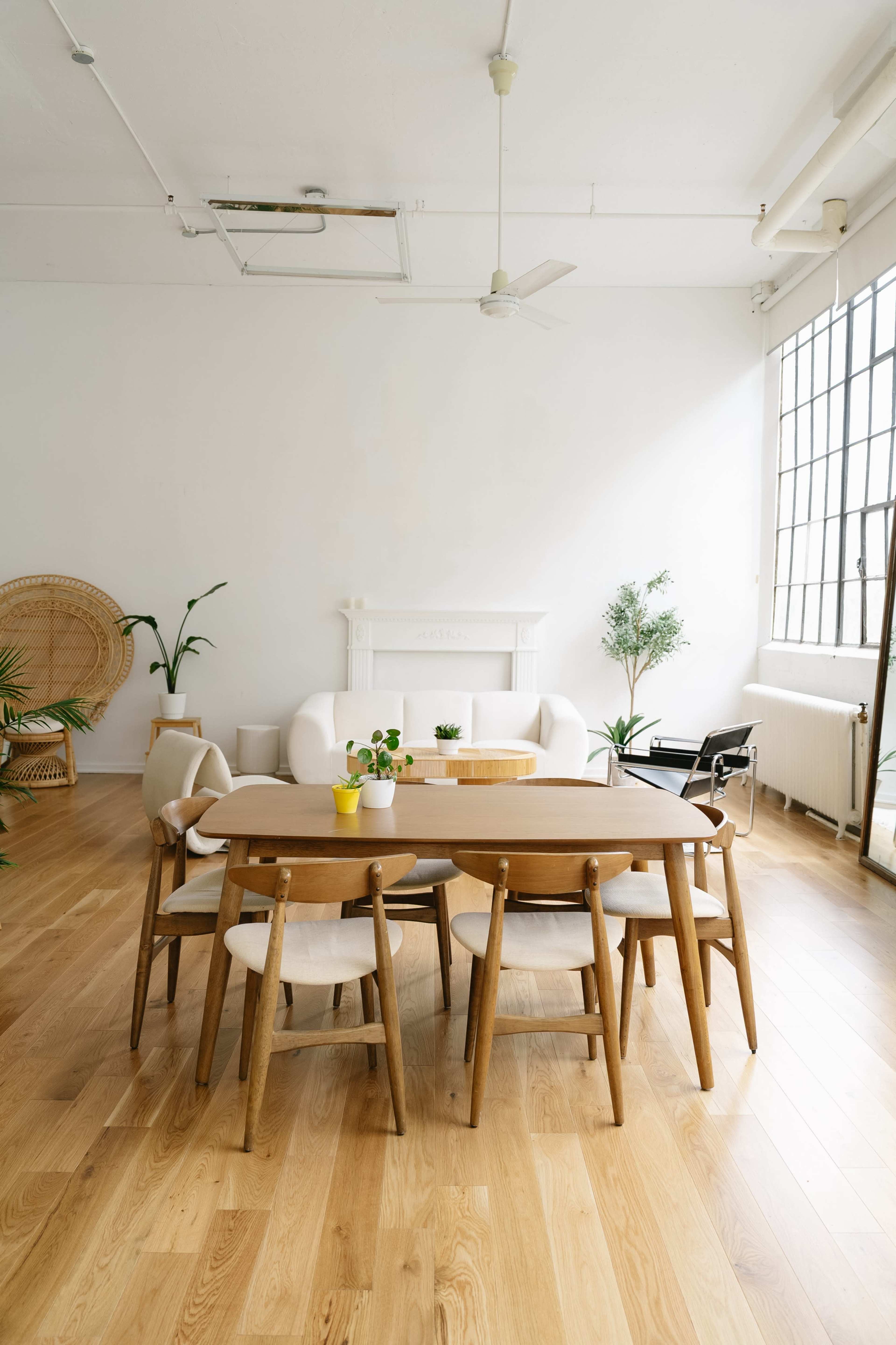 The image shows a spacious, minimalist dining area with a wooden table surrounded by six chairs, a white couch, and decorative plants against a light-colored wall.