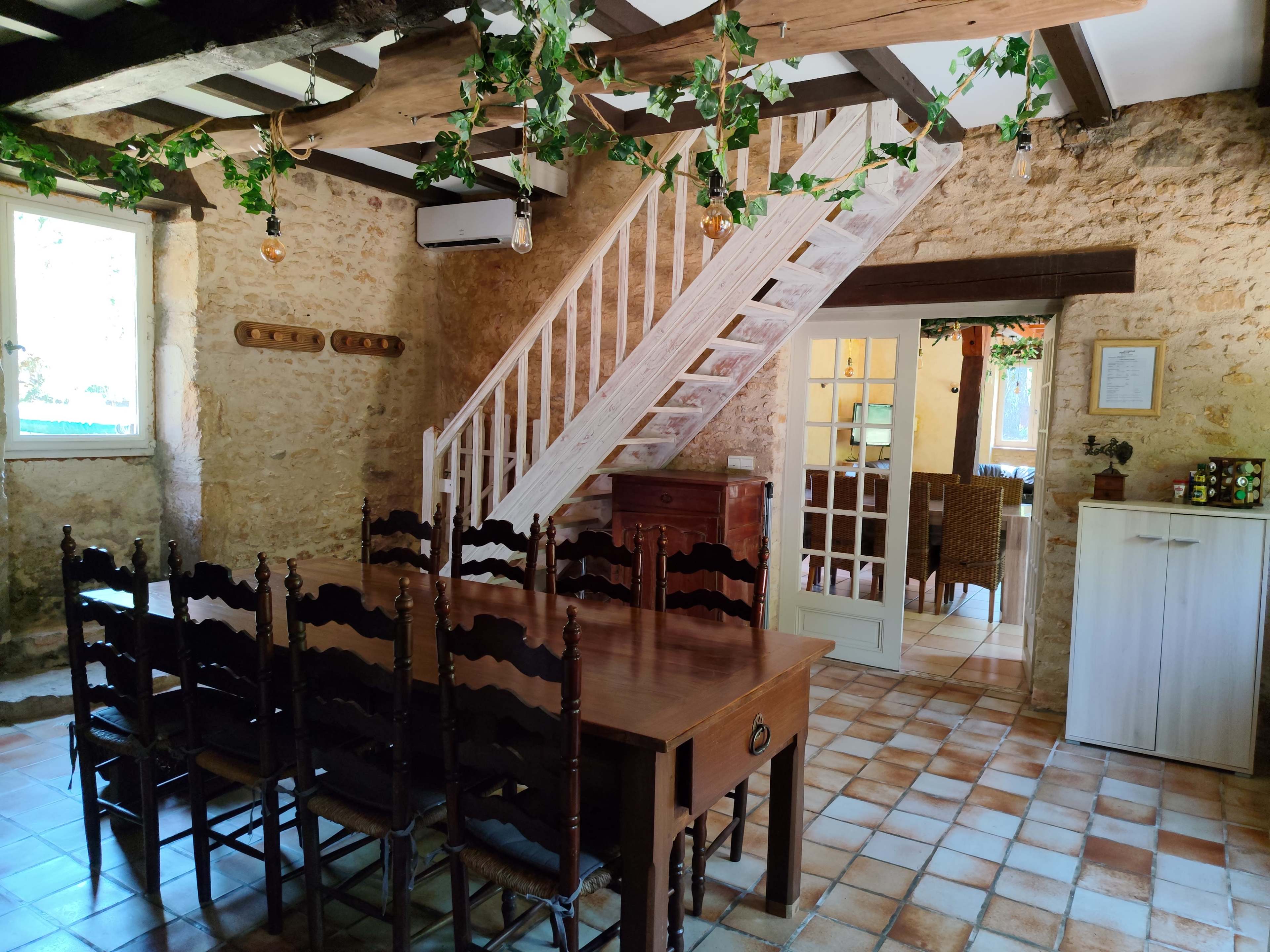 The image shows a rustic dining area with a wooden table and chairs, a staircase leading to an upper floor, and stone walls decorated with vines.