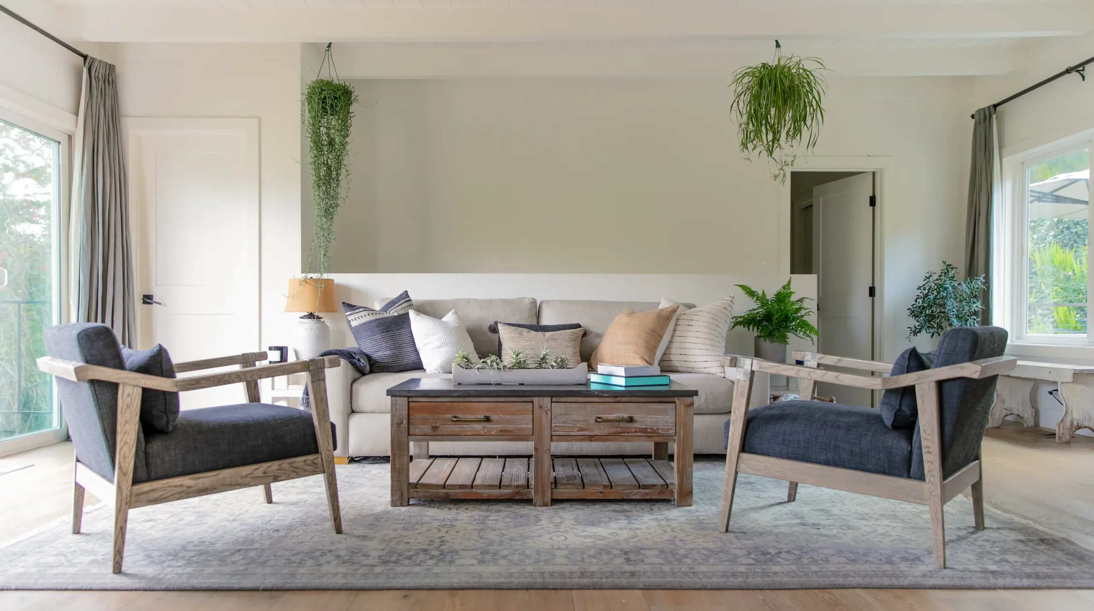 A living room with a beige sofa, two wooden armchairs, a coffee table made of reclaimed wood, and plants hanging from the ceiling.