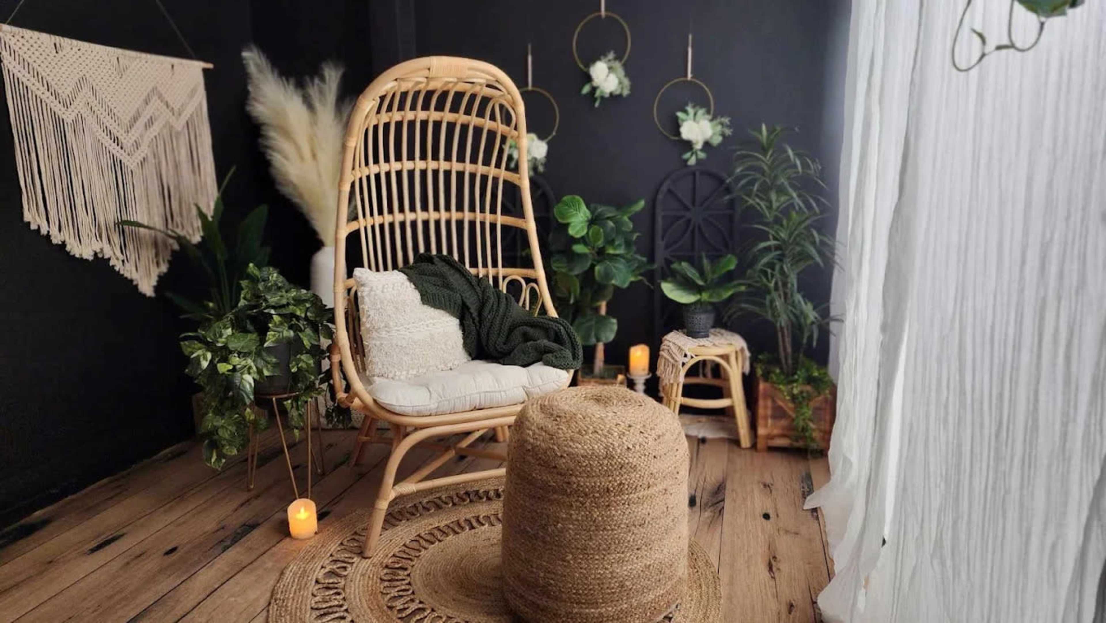 The image features a cozy indoor seating area with a rattan chair, a round woven pouf, and various indoor plants against a dark wall.