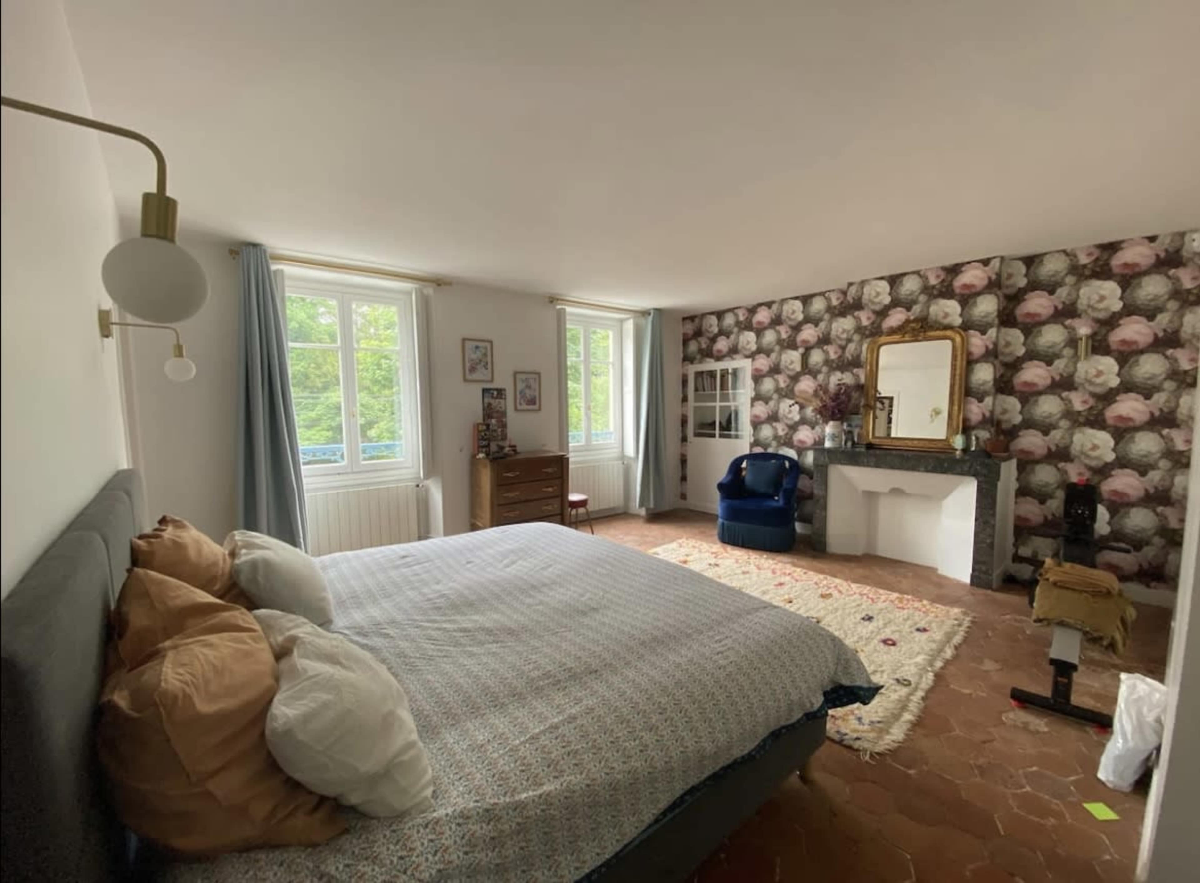 A bright bedroom featuring a large bed, a blue armchair, floral wallpaper, and a mirror above a fireplace.
