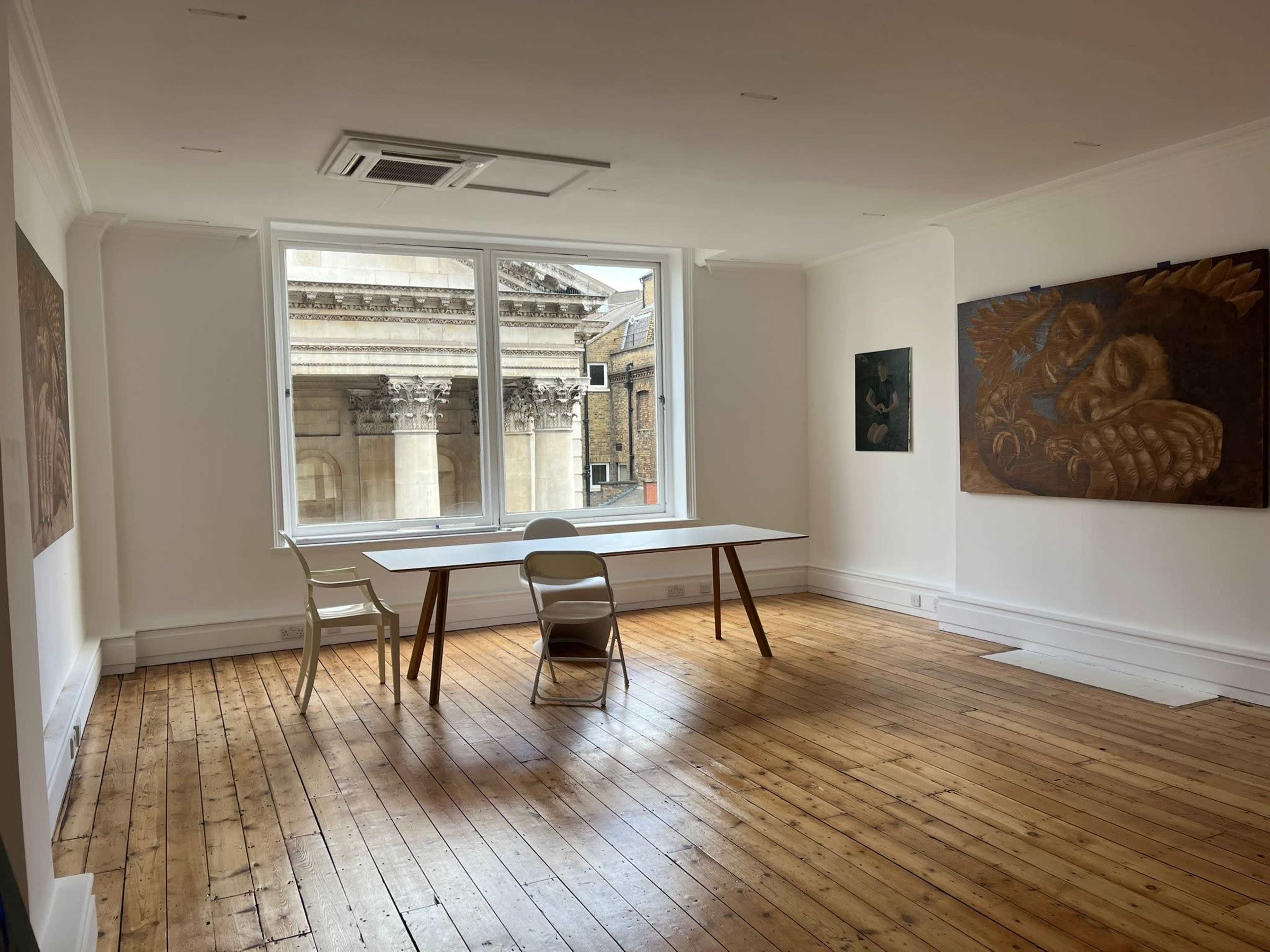 A bright, empty room with wooden floors, a large window facing a brick building, and a simple table with a chair.