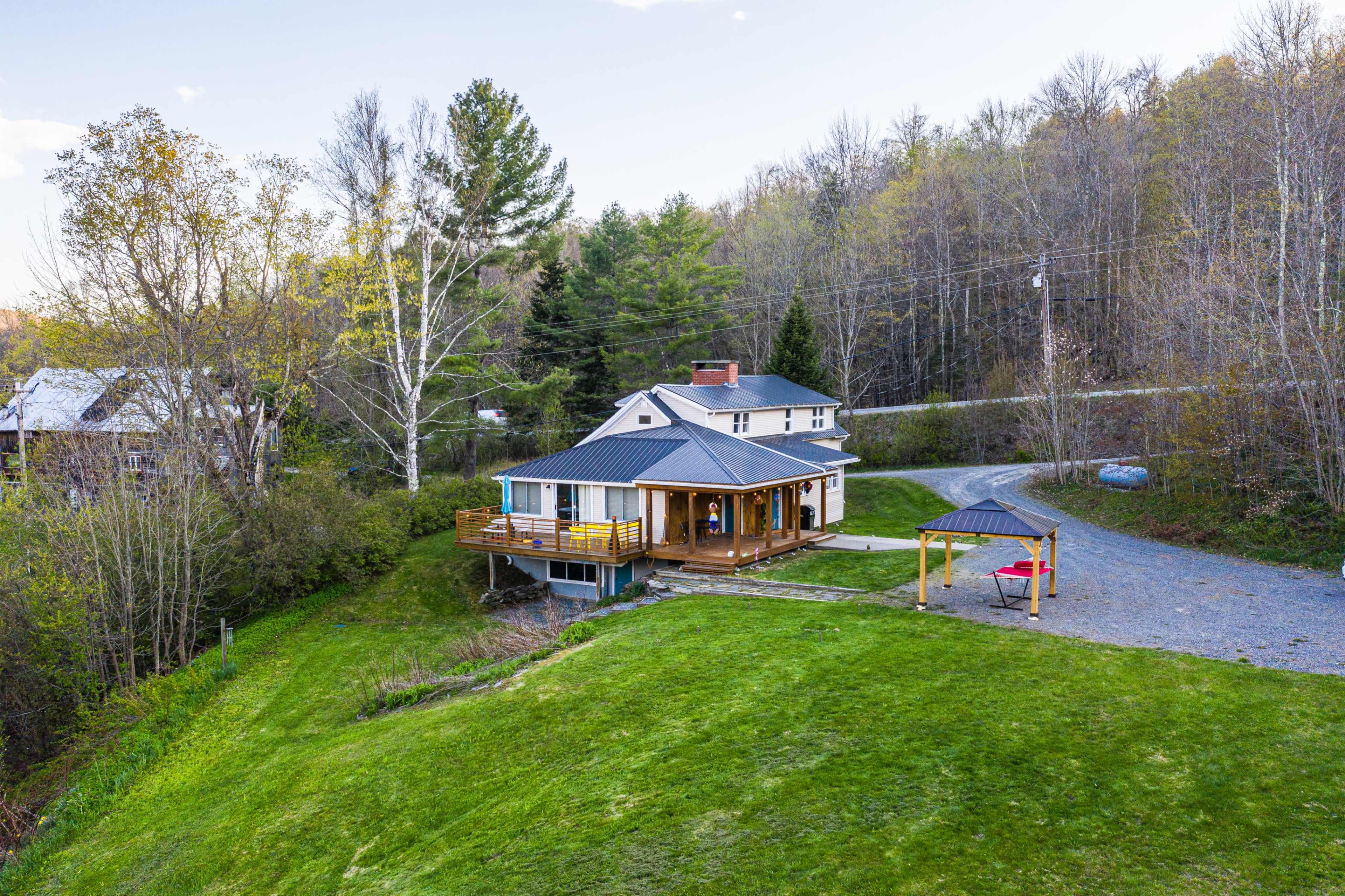 A two-story house with a wooden deck overlooks a grassy slope and is surrounded by trees.