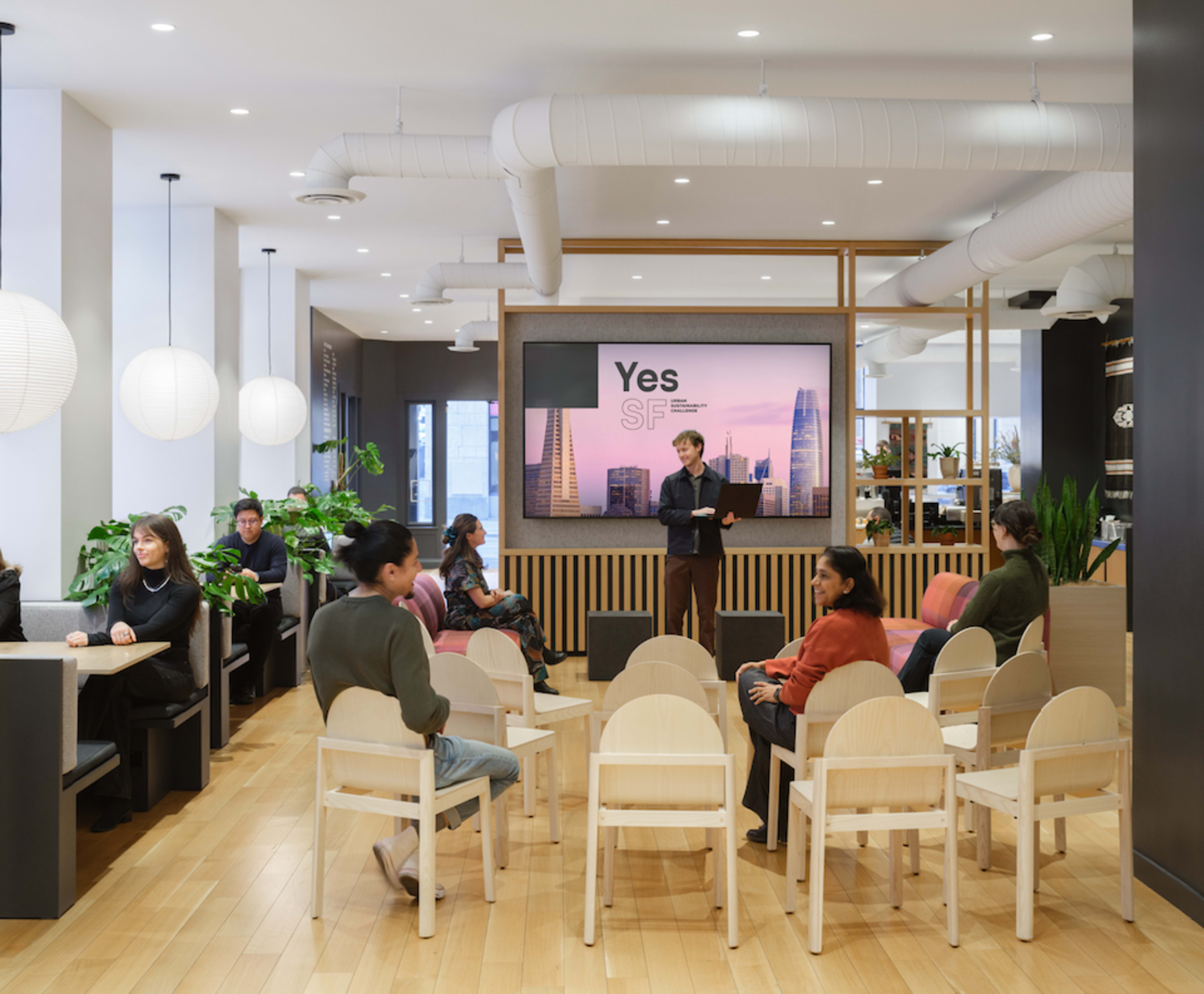 A modern meeting space features a speaker in front of a large screen, with several attendees seated in minimalist chairs around the room.