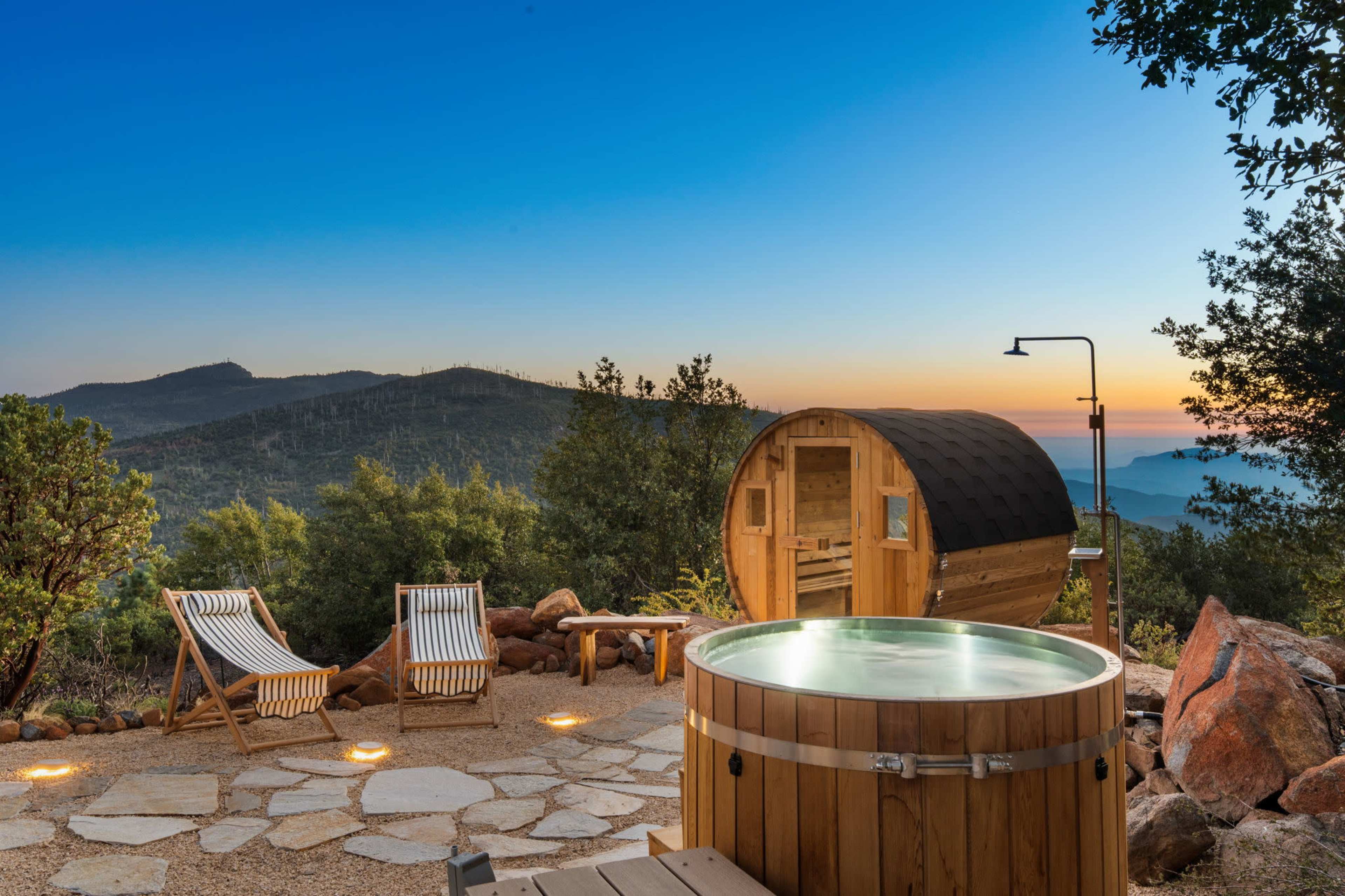 The image features a wooden hot tub and a small cabin set against a mountainous landscape at dusk, with two lounge chairs in the foreground.