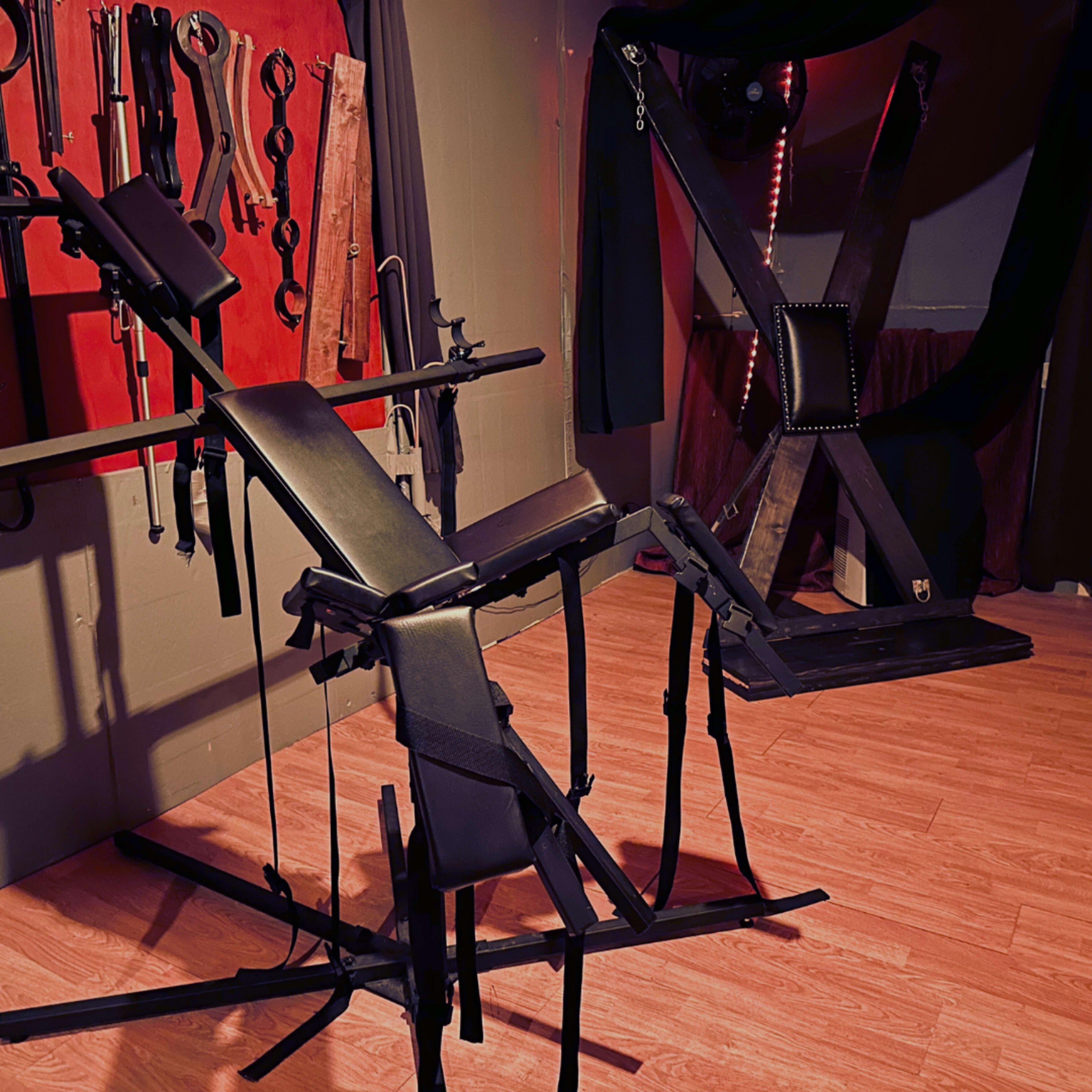 The image shows a dimly lit room featuring various BDSM equipment, including a harness and an X-frame restraint structure.