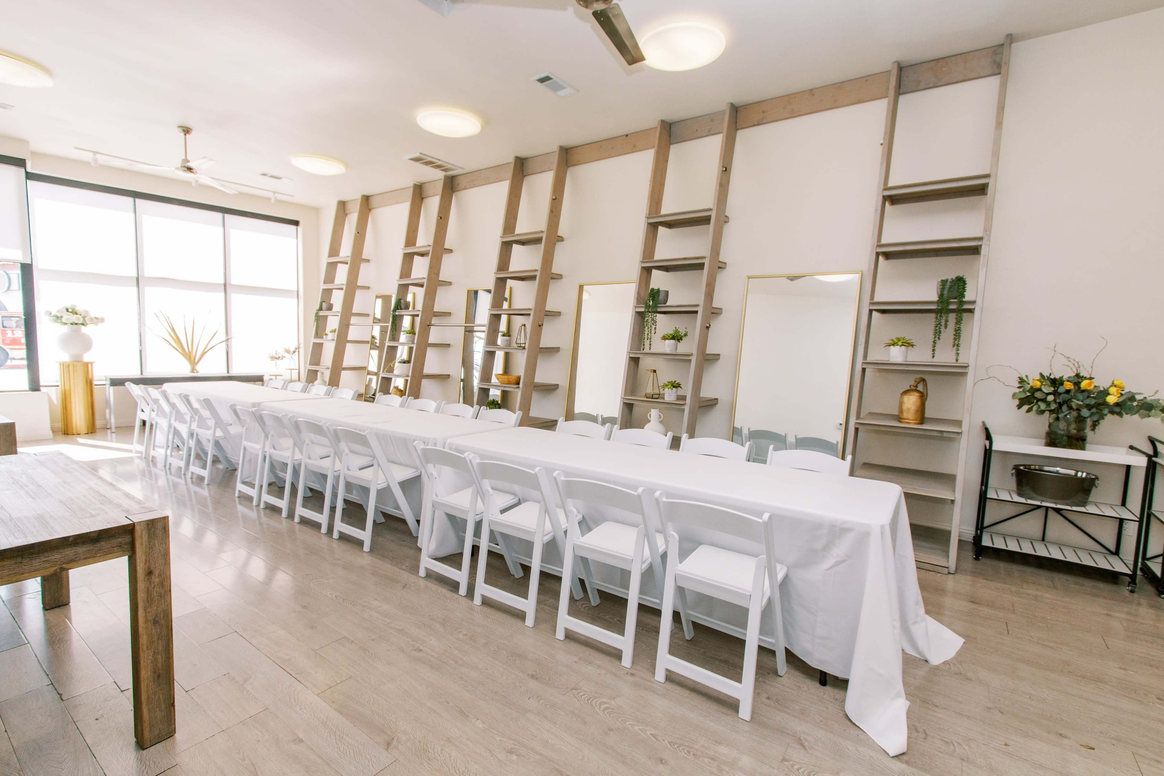 A long table is set with white tablecloths and surrounded by white folding chairs in a well-lit room featuring wooden shelves on the walls.