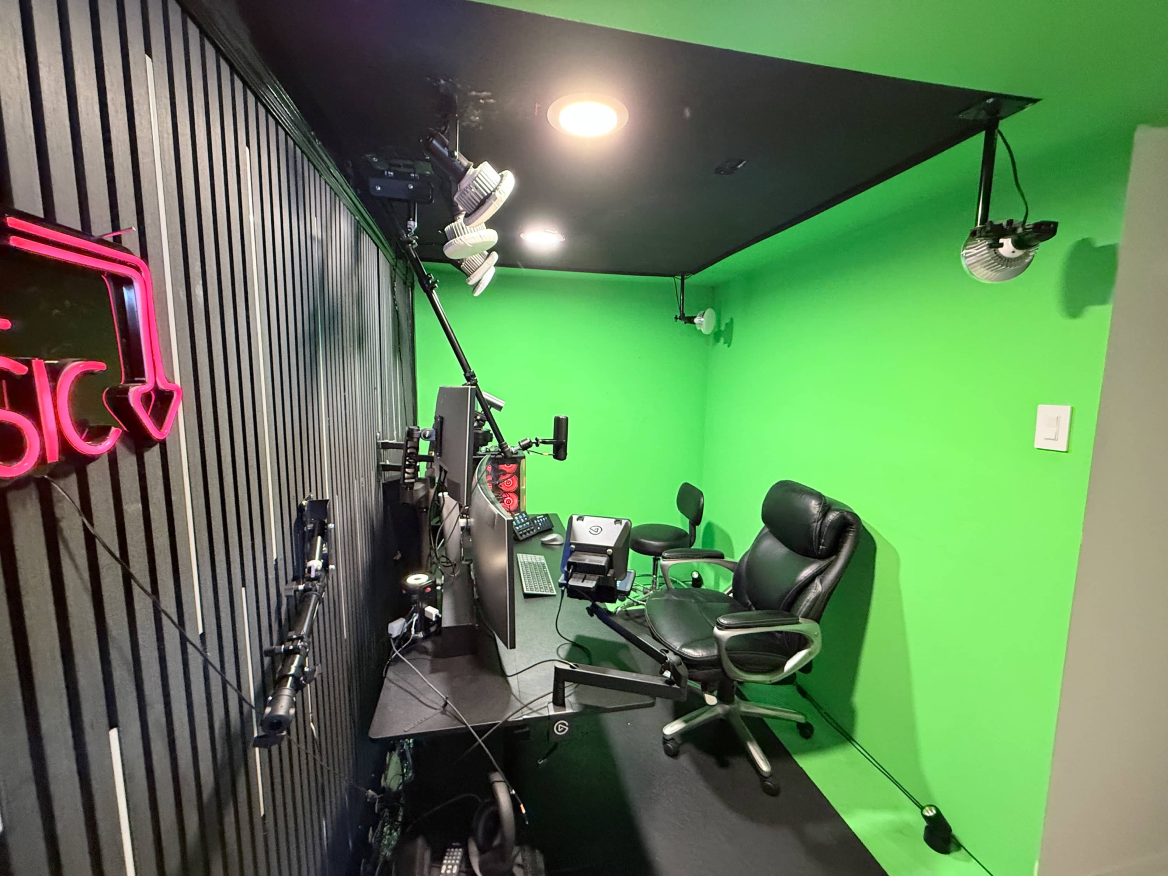 The image shows a compact recording studio with a green backdrop, a desk with a computer and microphone, and two office chairs.
