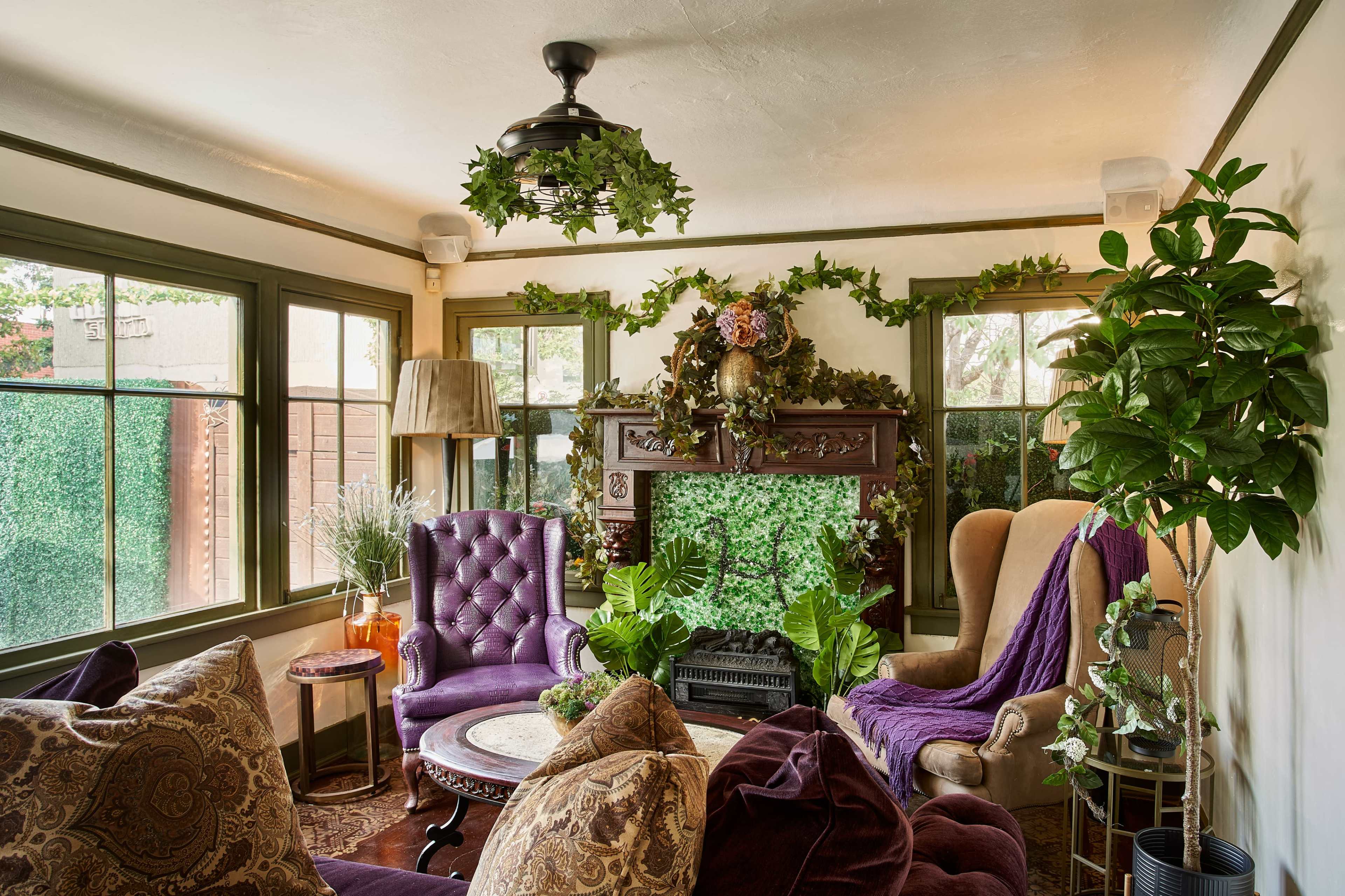The scene features a cozy living room with purple upholstered chairs, a decorative fireplace adorned with plants, and large windows allowing natural light.