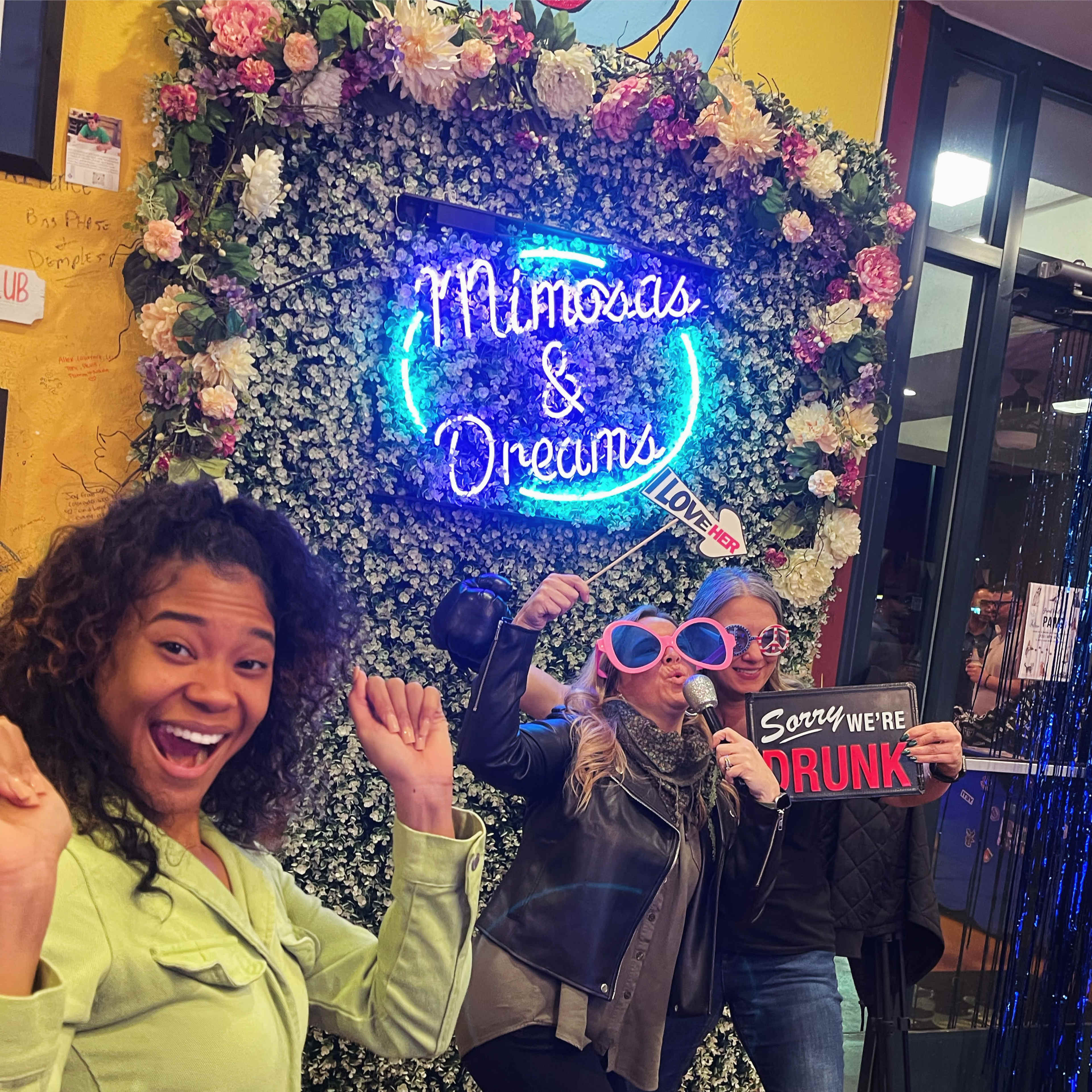 Three people pose playfully in front of a floral backdrop with a neon sign that reads "Mimosas & Dreams."