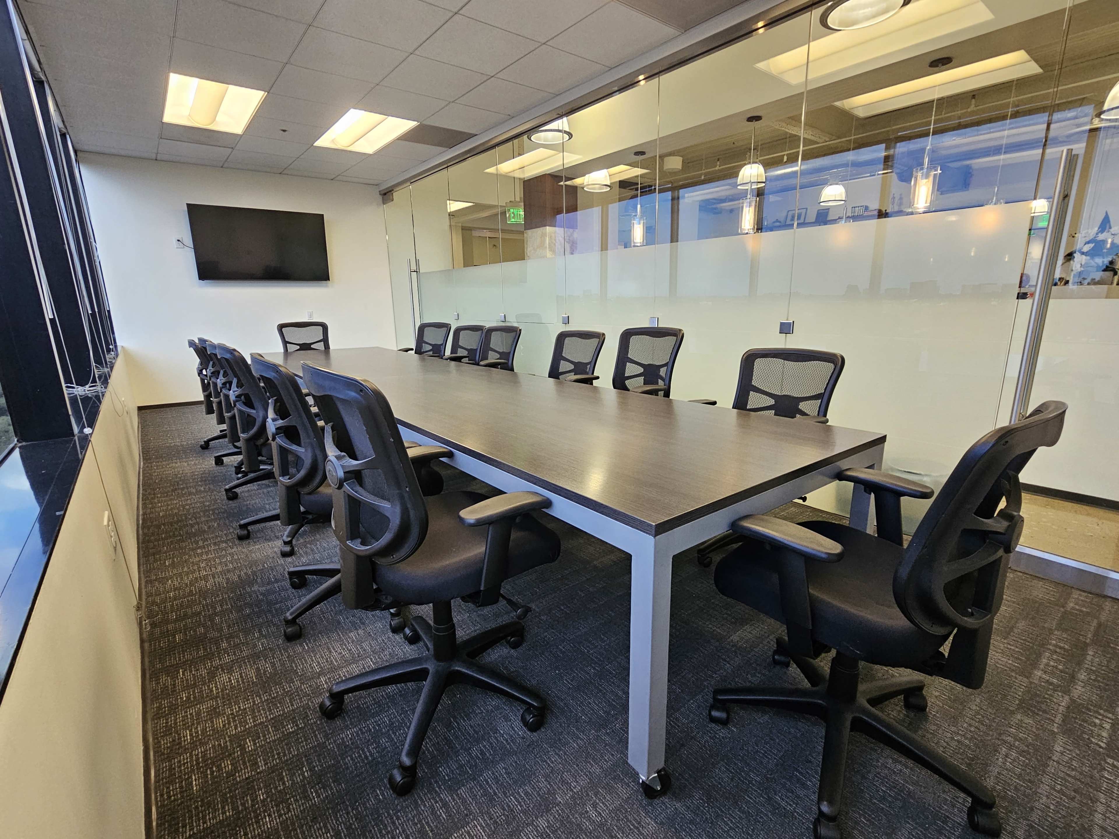 A long, rectangular conference table is surrounded by ergonomic office chairs in a modern glass-walled meeting room.