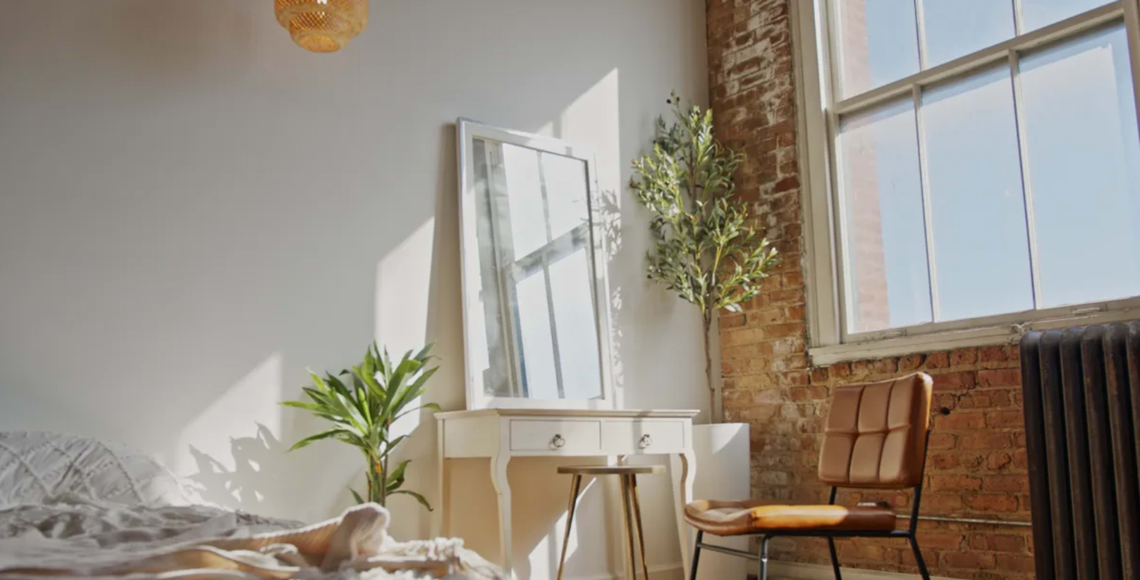 A bright bedroom features a large mirror on a white dresser, a plant, a wooden stool, and a leather chair near a window with natural light illuminating the space.