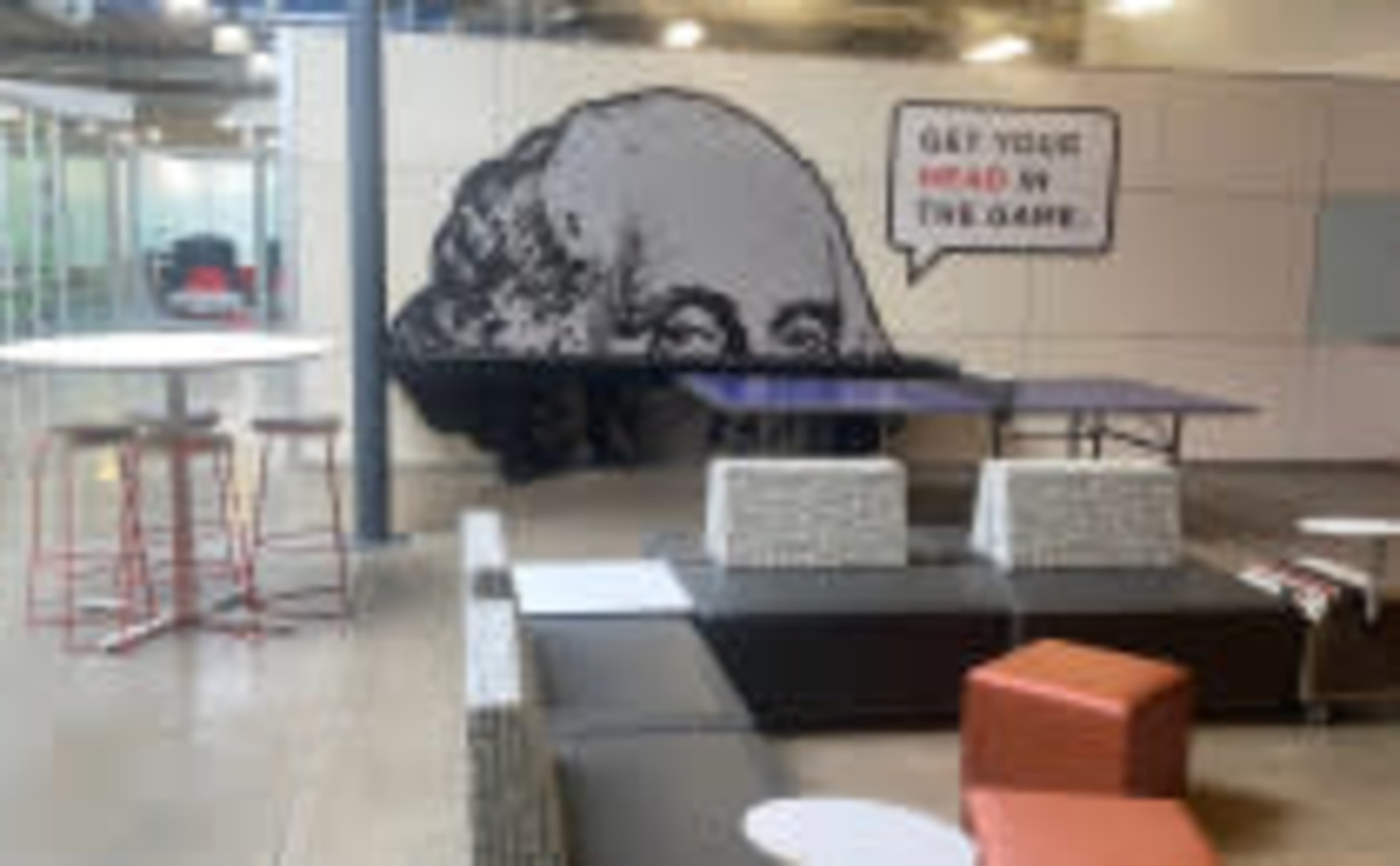 A modern workspace with a large mural of a man's head and a motivational phrase on the wall, along with various seating areas and tables.