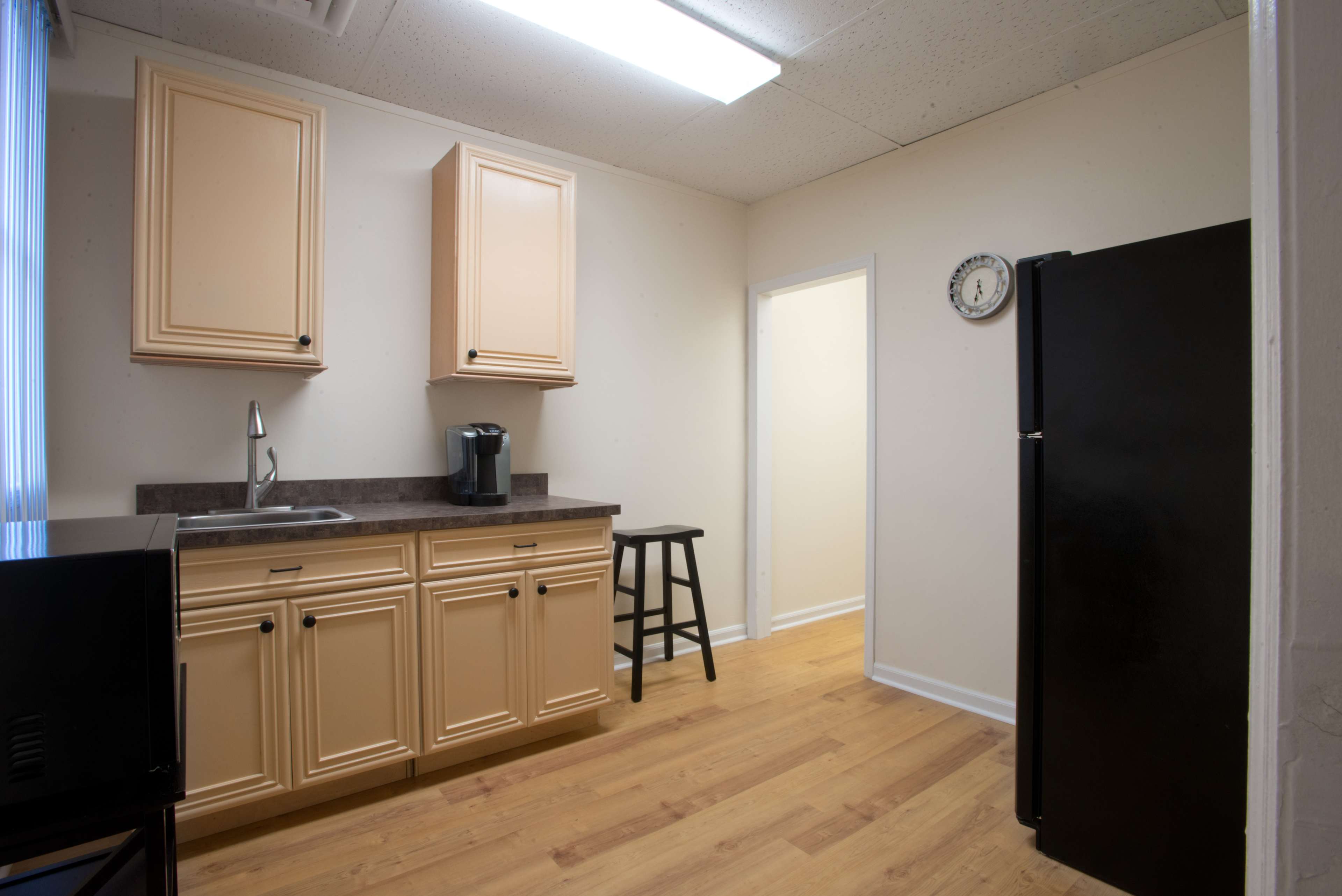 A small kitchen with light-colored cabinets, a dark countertop, a coffee maker, a black refrigerator, and a single stool.