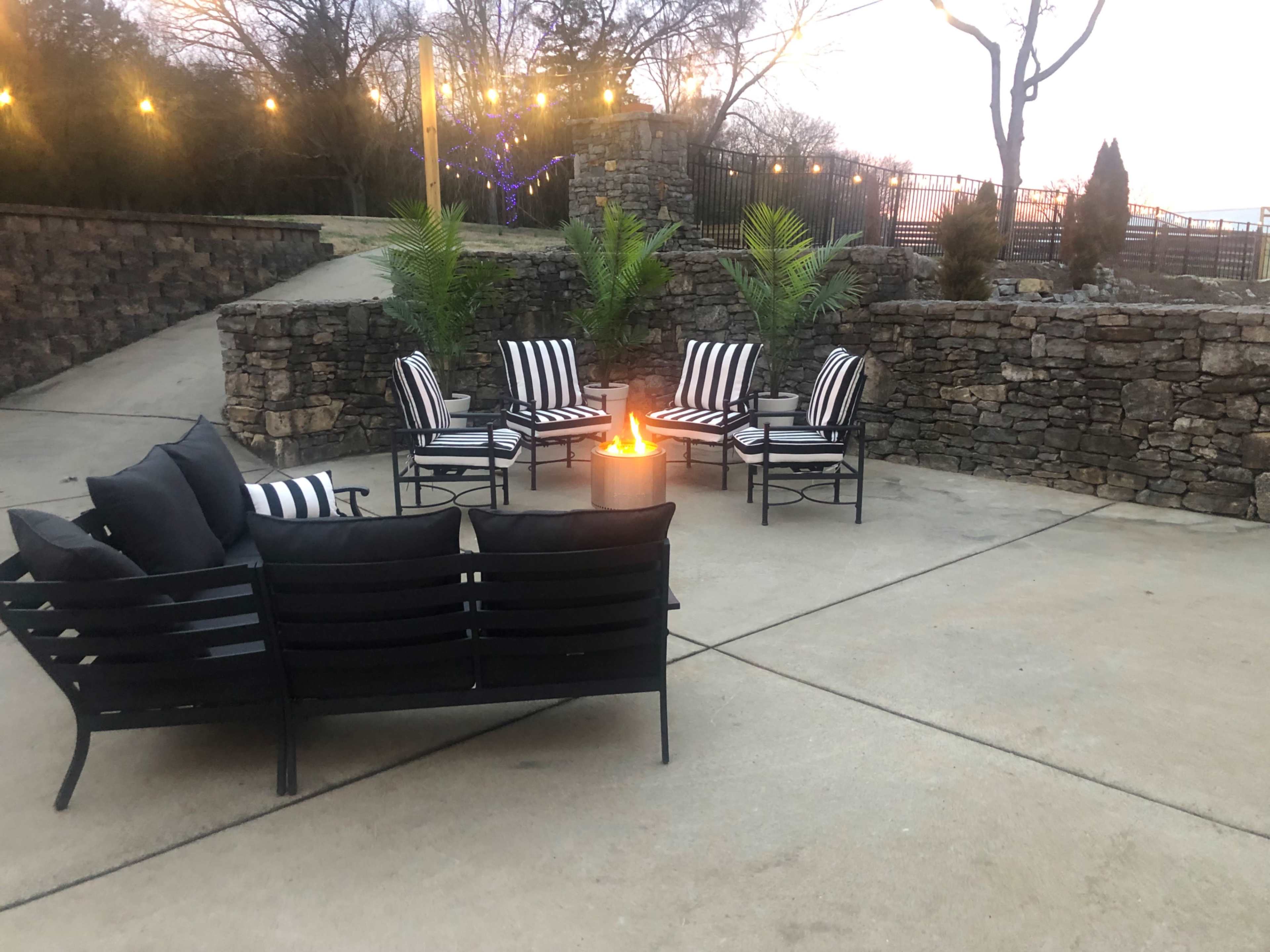 A circular seating arrangement with black and white striped chairs surrounds a fire pit on a stone patio, illuminated by string lights in the background.