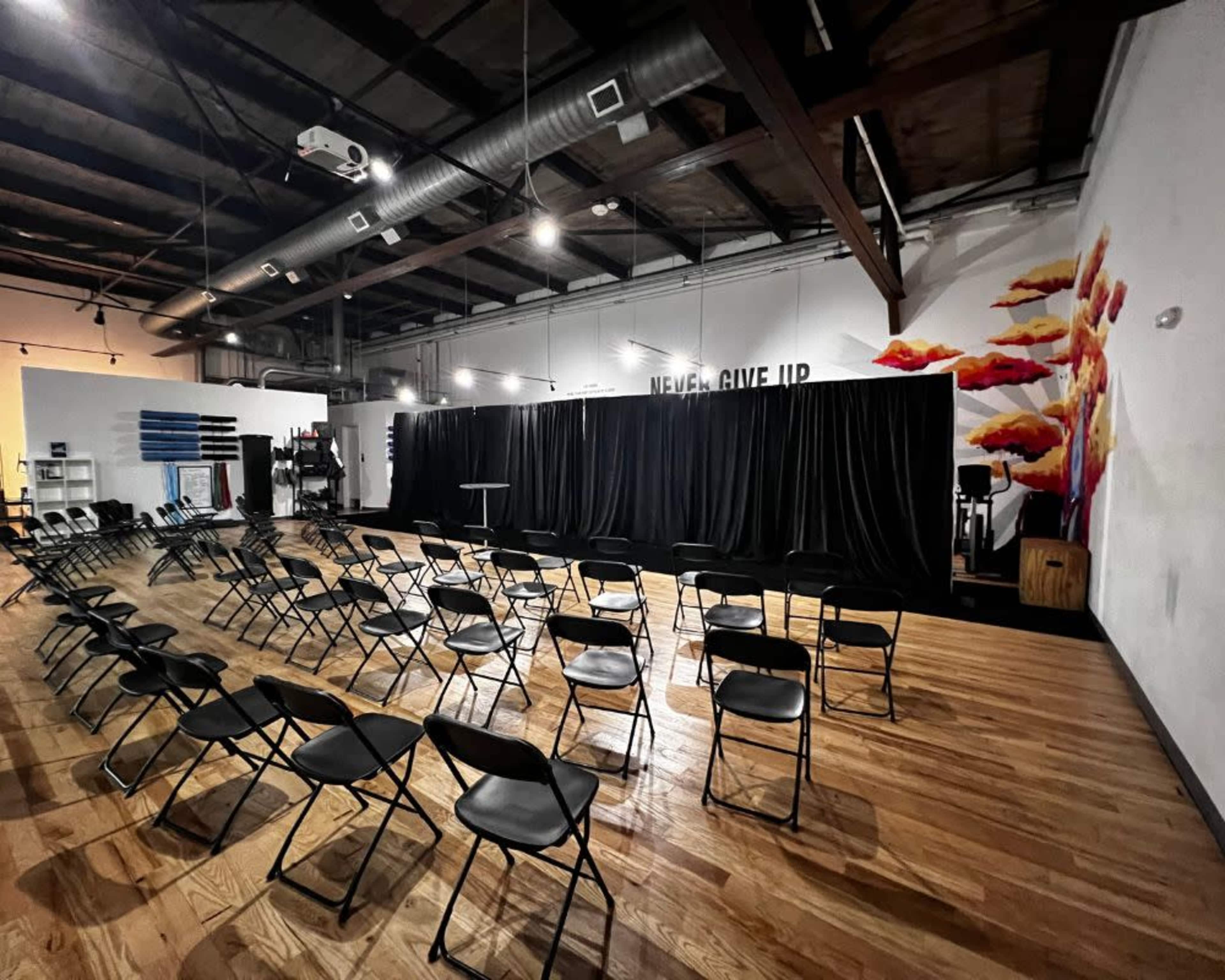 A spacious room set up with rows of black chairs facing a black curtain stage, with motivational text on the wall and a colorful mural.