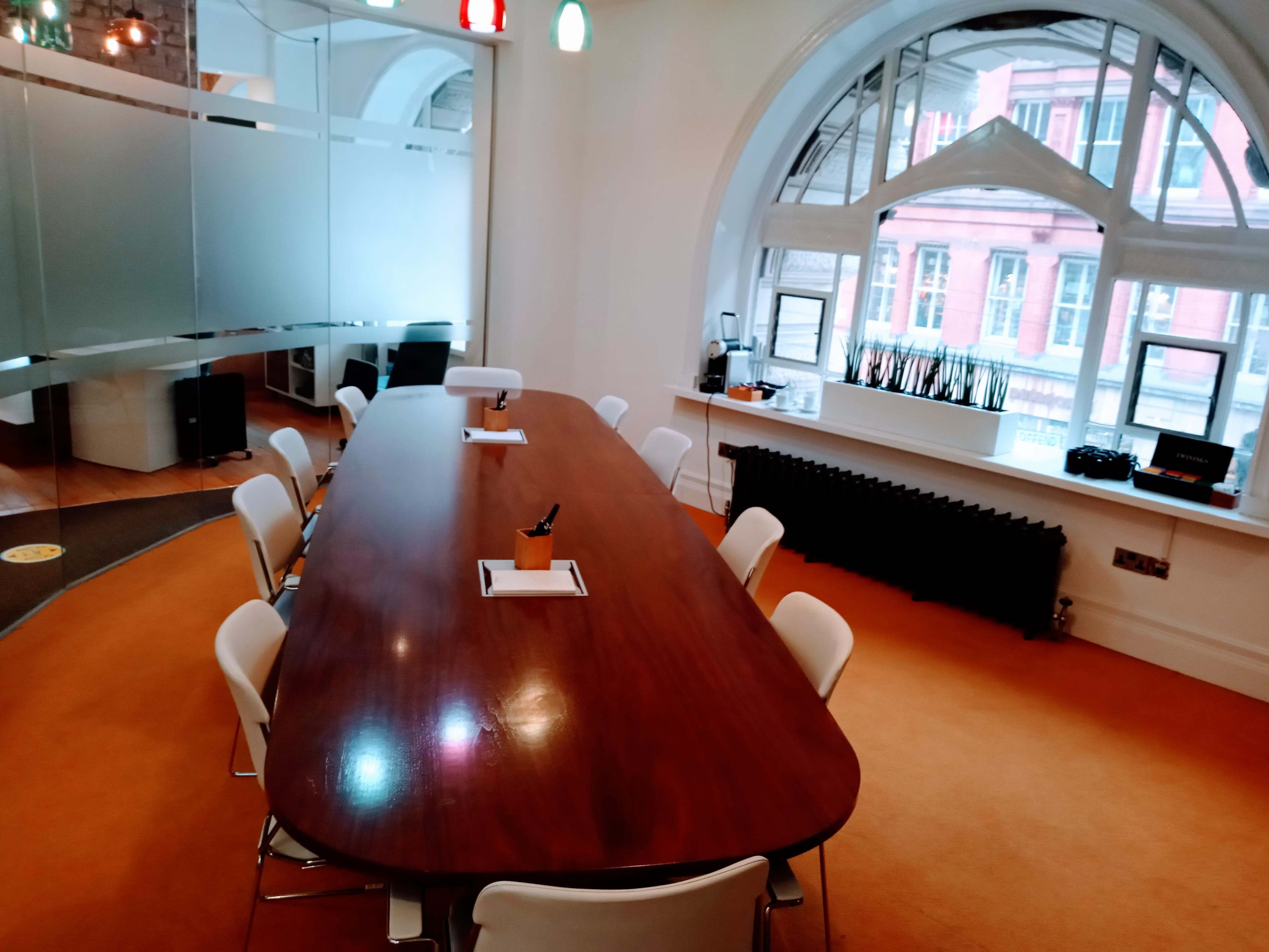 A large, oval wooden conference table is surrounded by white chairs in a brightly lit meeting room with a large window showing a view of a building exterior.