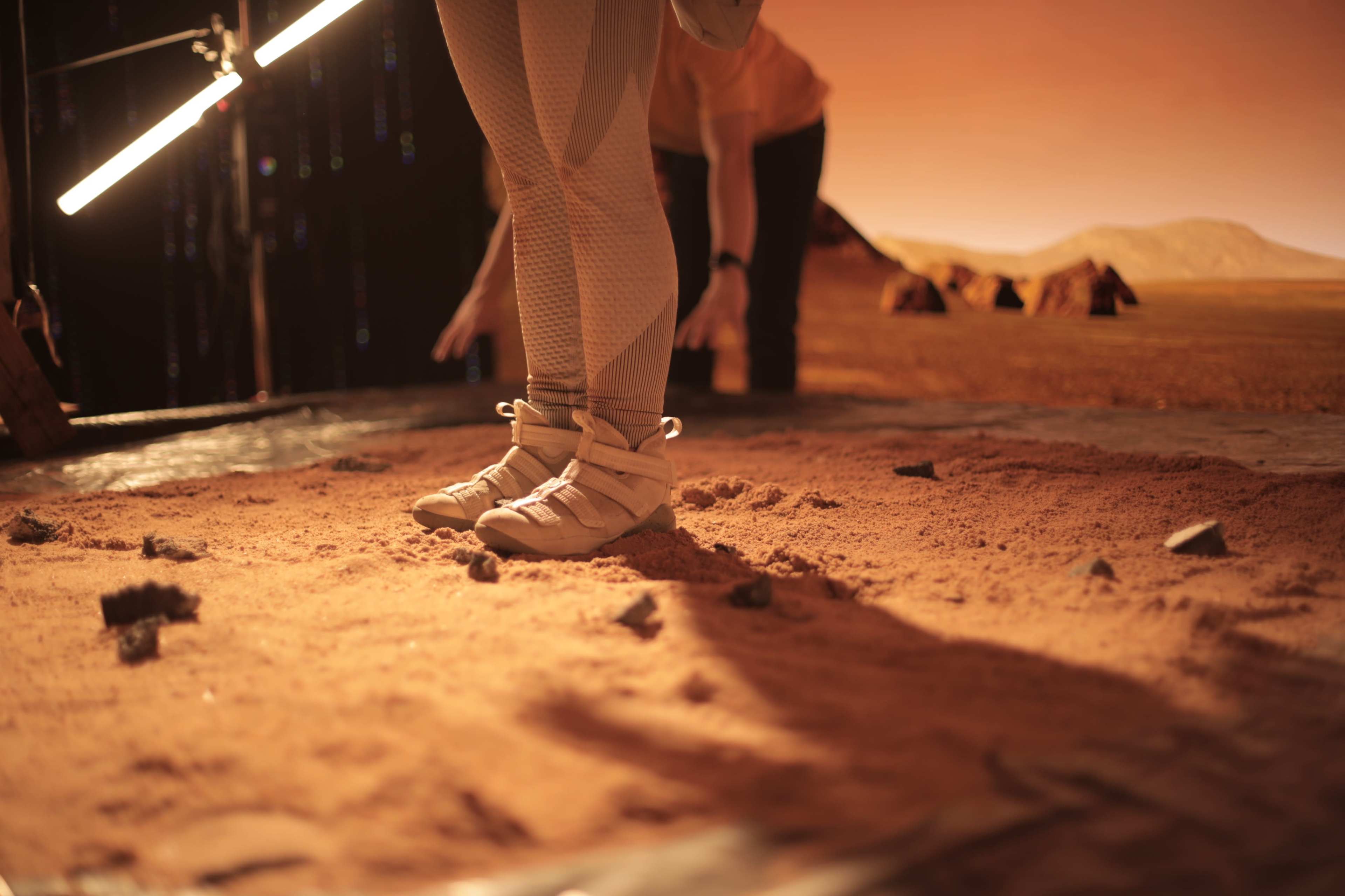 A person wearing light-colored footwear standing on a surface that resembles Martian terrain, with a backdrop of rocky formations and an orange sky.