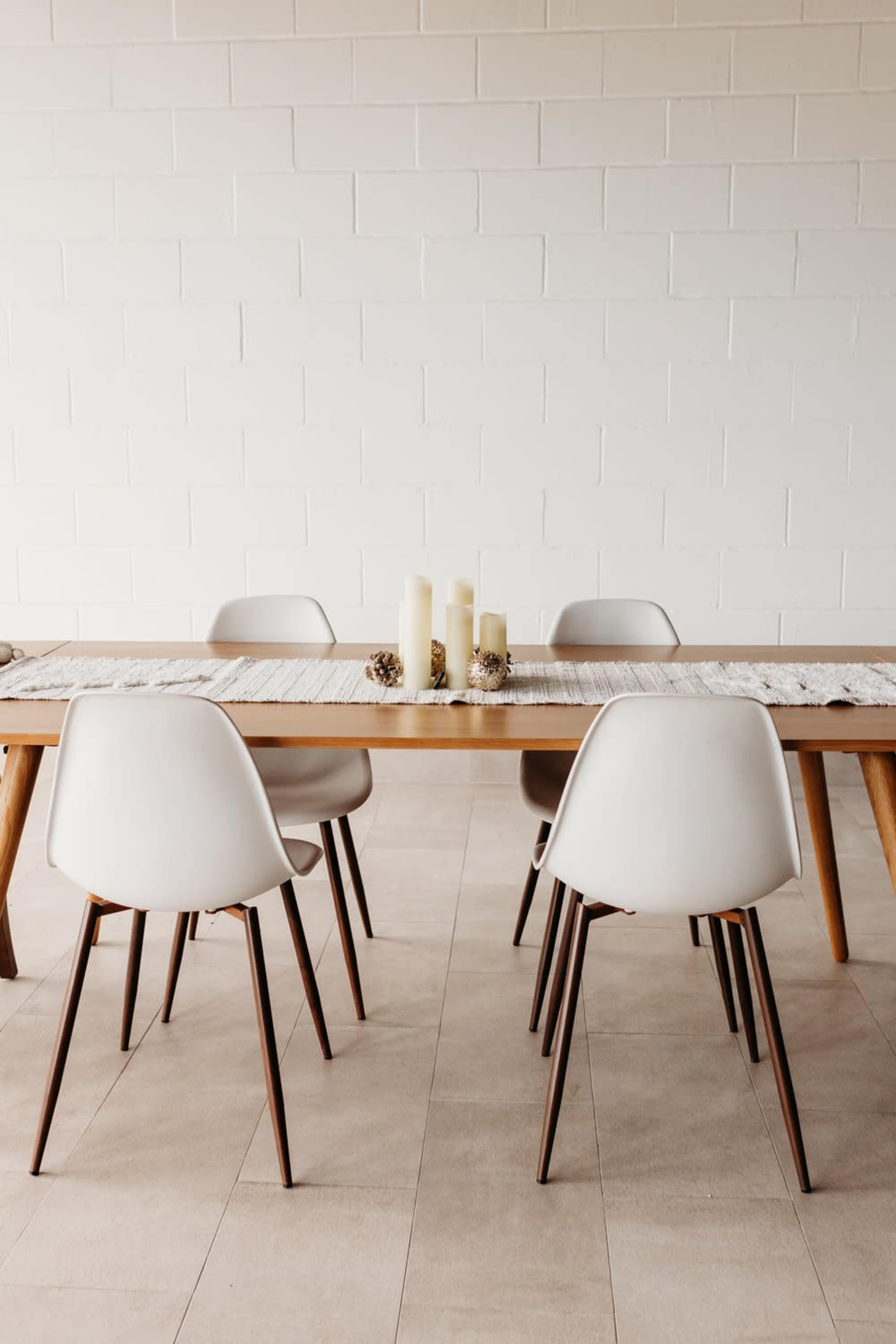 A wooden dining table is set with white candles and surrounded by four light-colored chairs on a tiled floor.