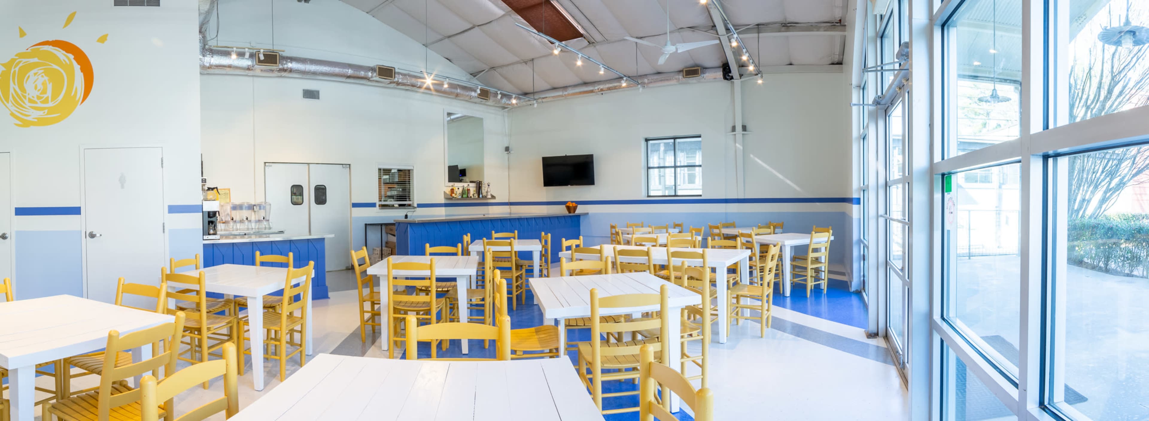 The image shows a bright and spacious dining area with yellow chairs and white tables, featuring large windows and a serving counter in the background.