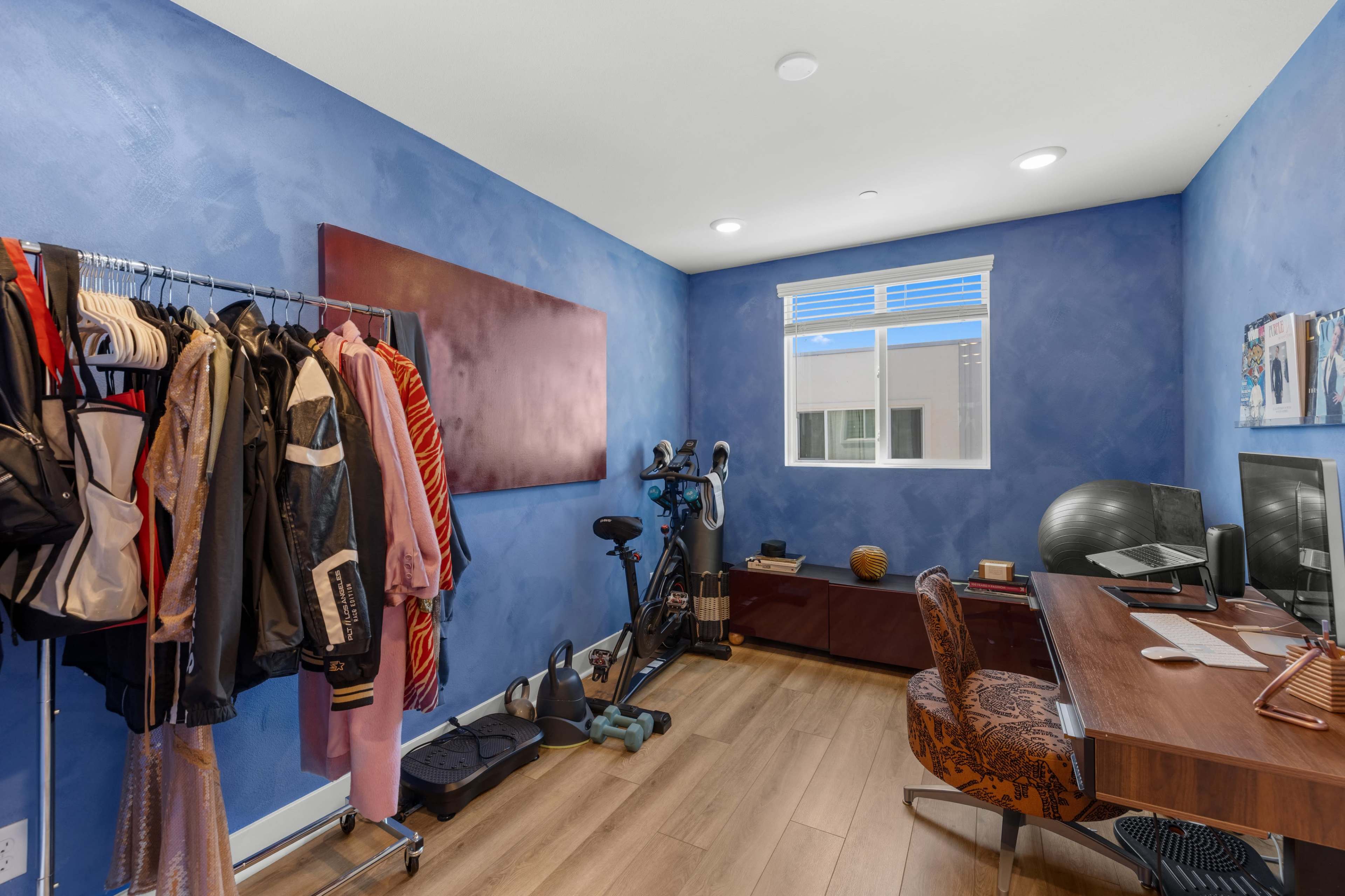The image shows a room with blue walls furnished with a desk, a computer setup, a stationary bike, exercise equipment, and a clothing rack holding several garments.