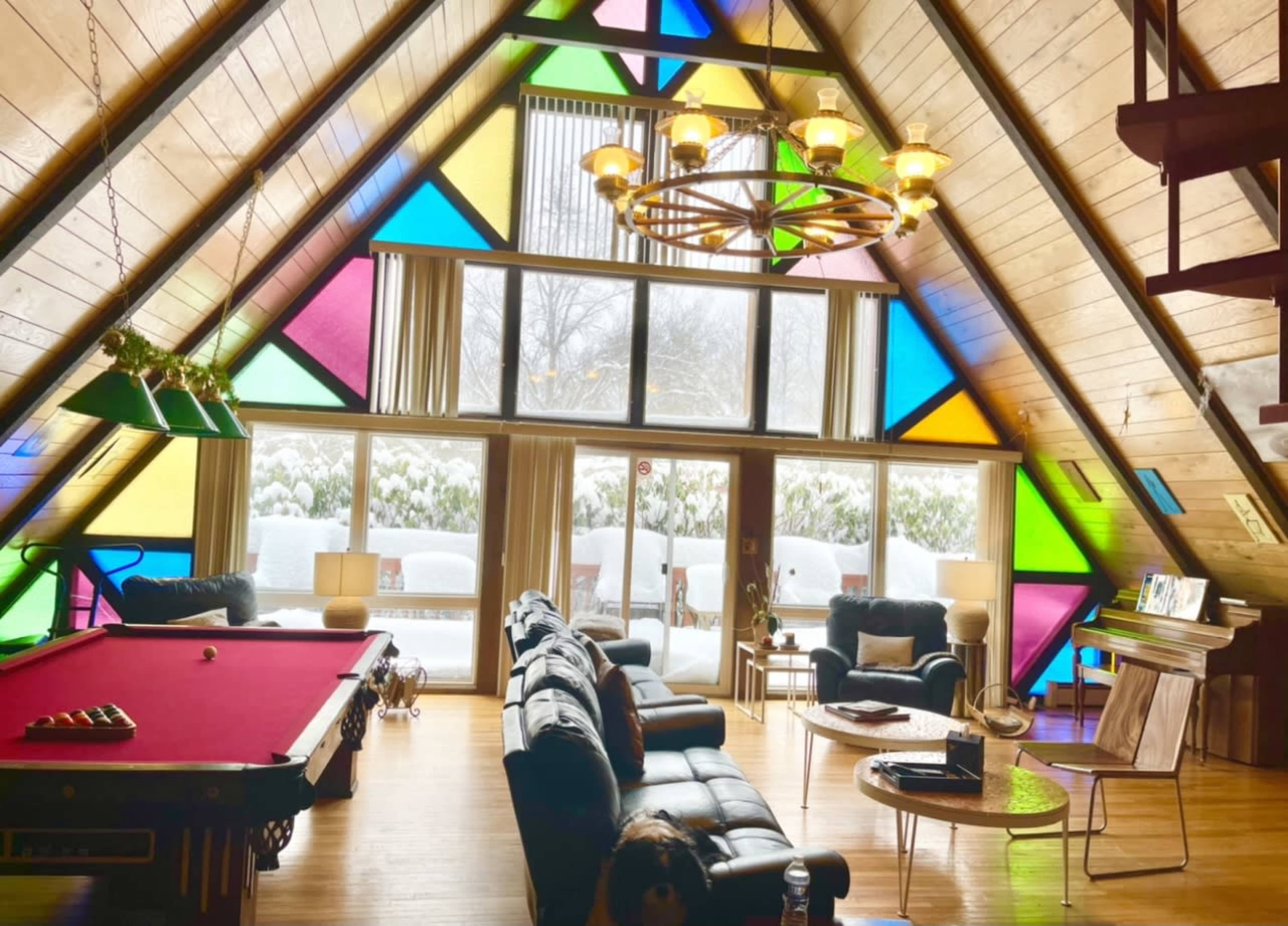 The image shows a spacious A-frame living room with colorful stained glass windows, a pool table, and large windows overlooking a snowy landscape.