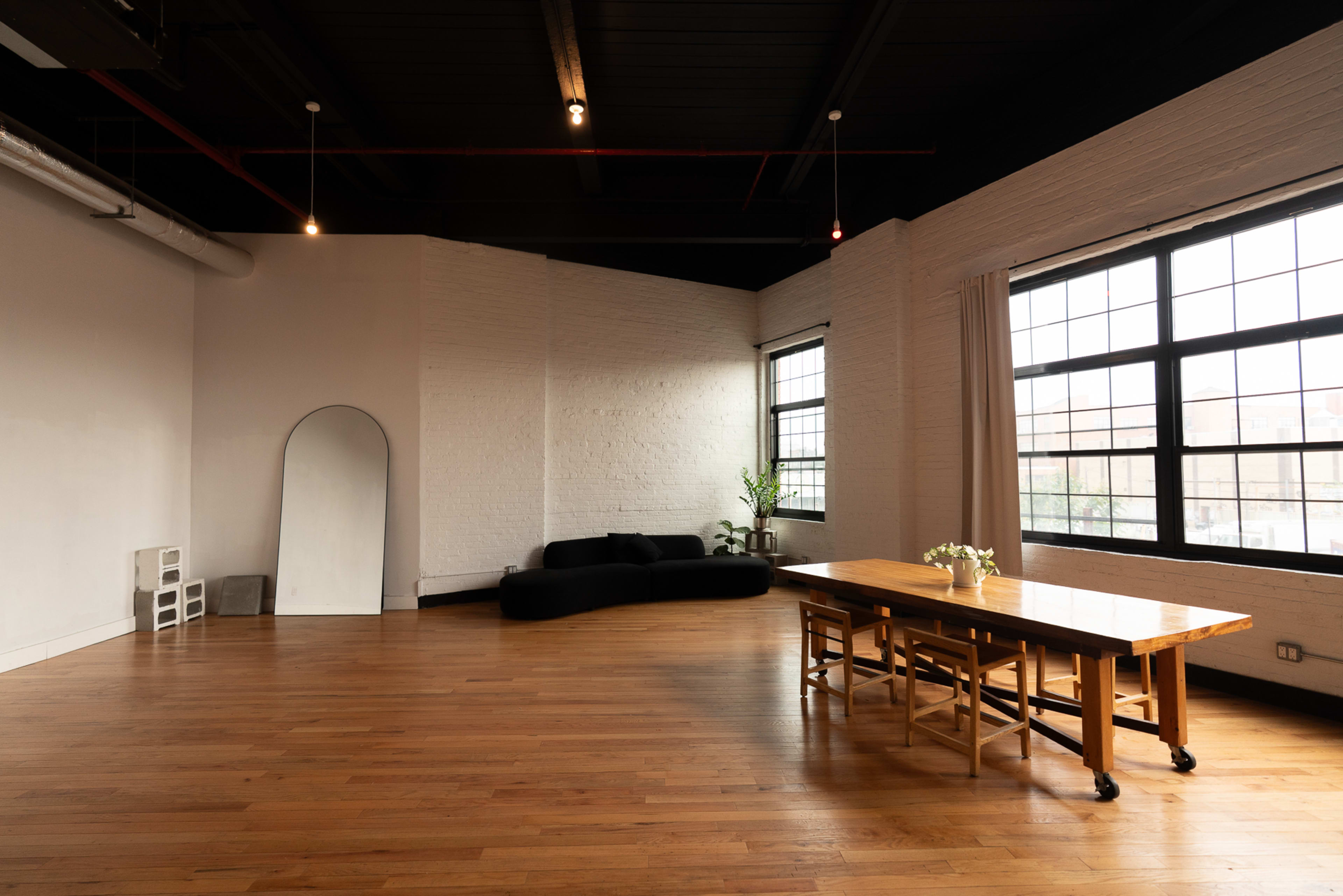 The image depicts a spacious, minimalist room featuring a large wooden dining table, a black sofa, and large windows that allow natural light to enter.