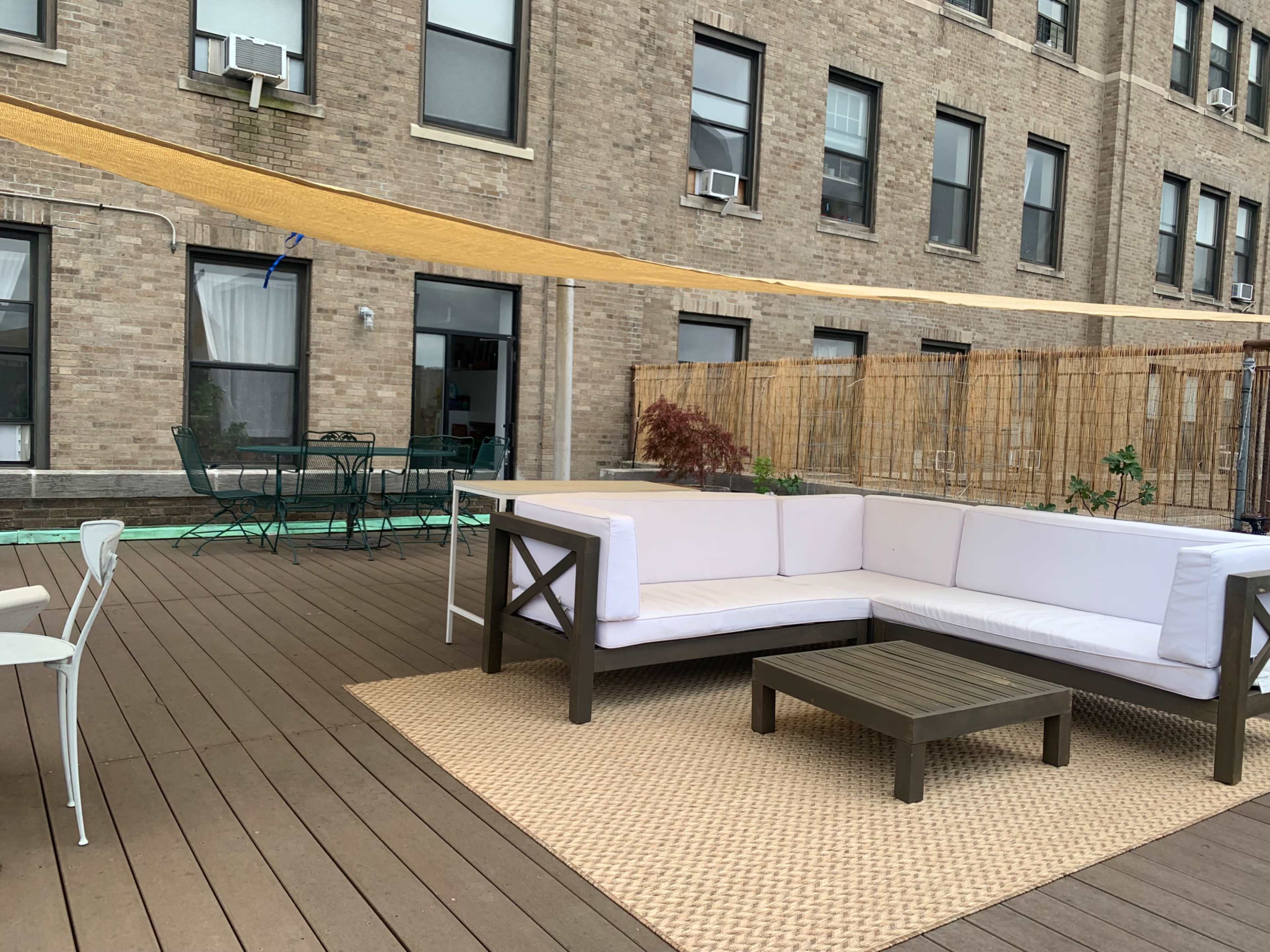 A patio features a white sectional couch and a low coffee table on wooden decking, surrounded by a brick building with several windows.