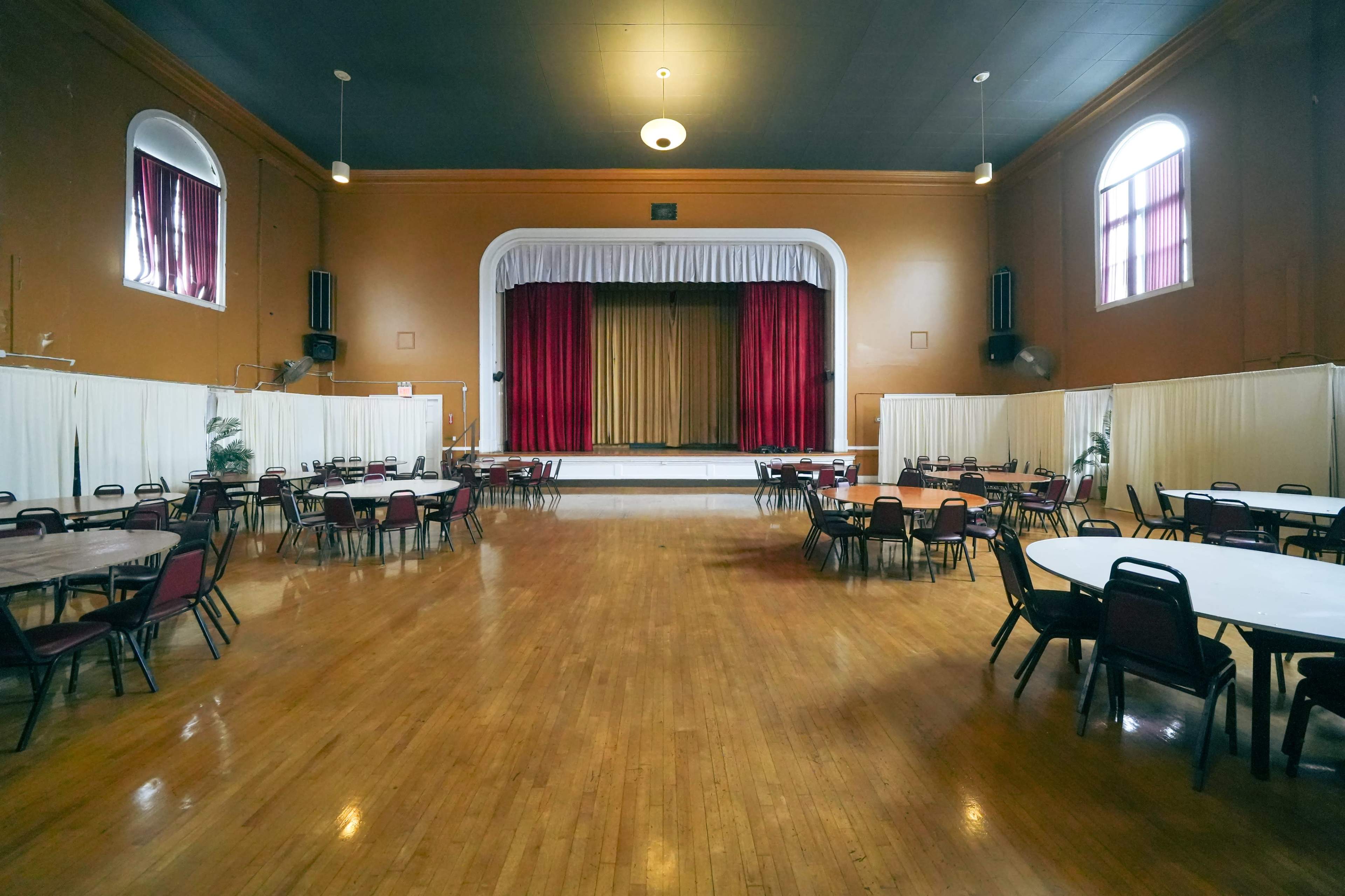 A large, empty hall features round tables and a stage with red curtains at the front.
