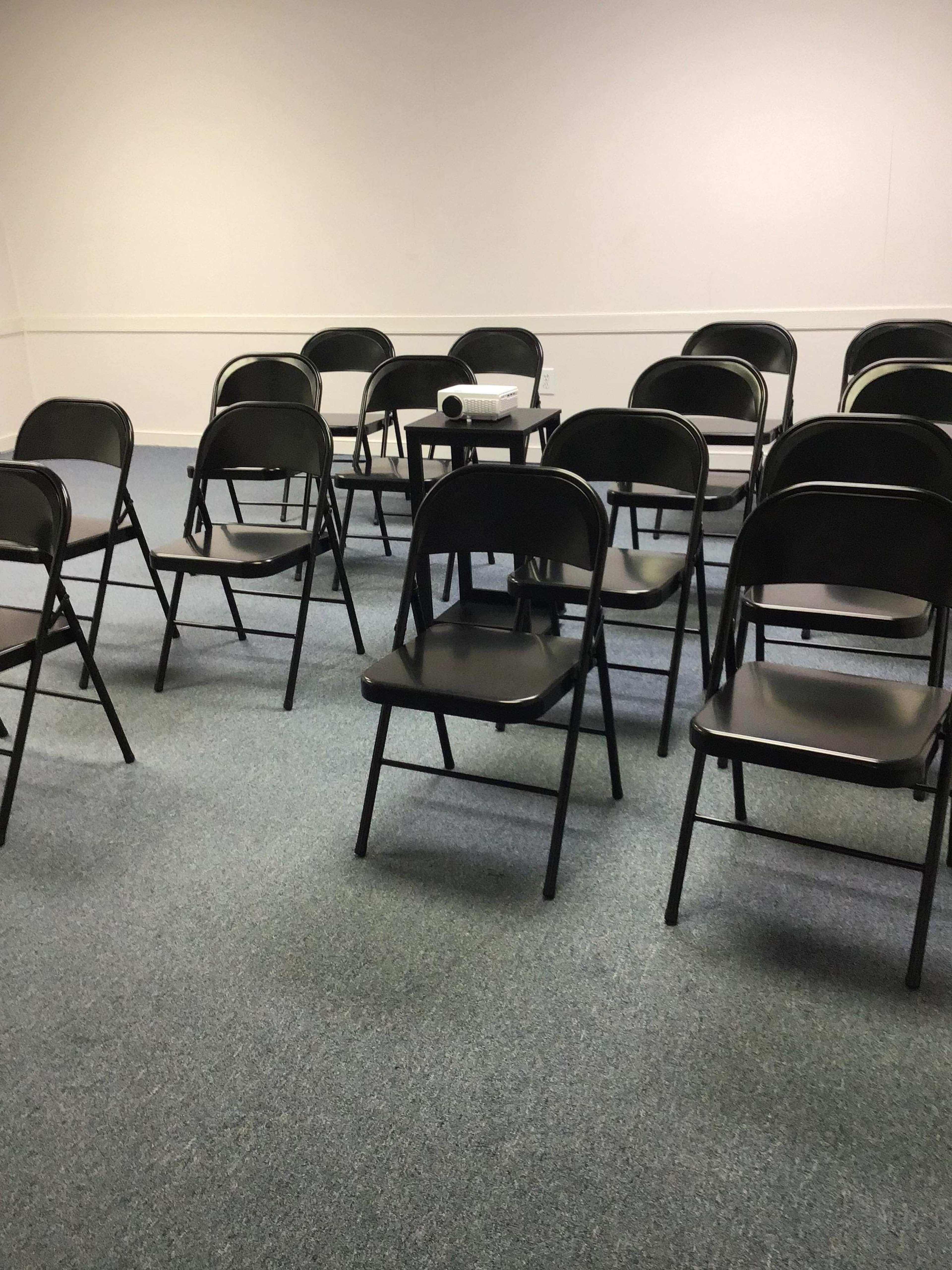 A room with several black folding chairs arranged around a central table featuring a projector.