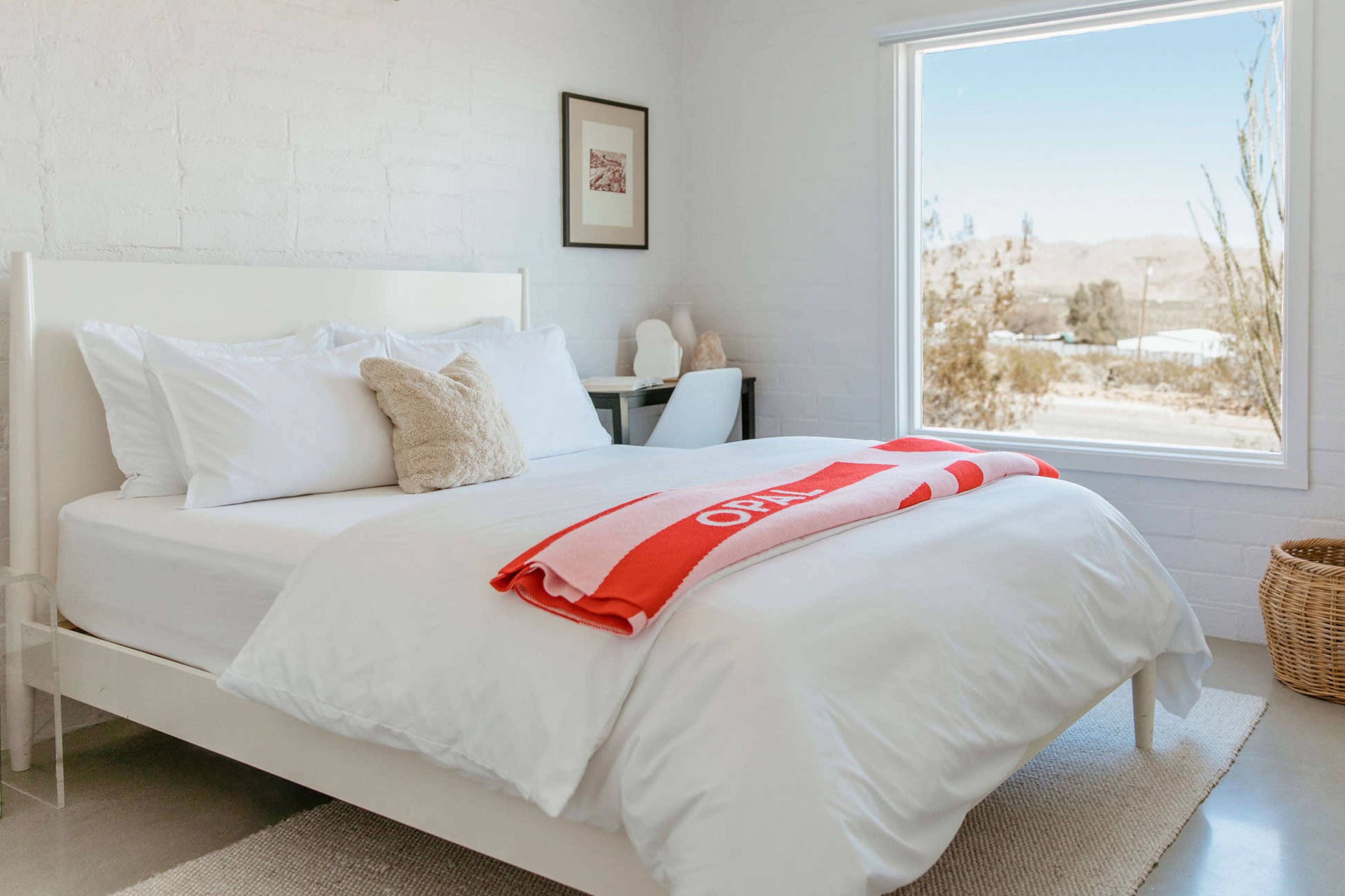 A neatly made bed with white linens and a red blanket is located in a bright room featuring a large window that offers a view of the outdoors.