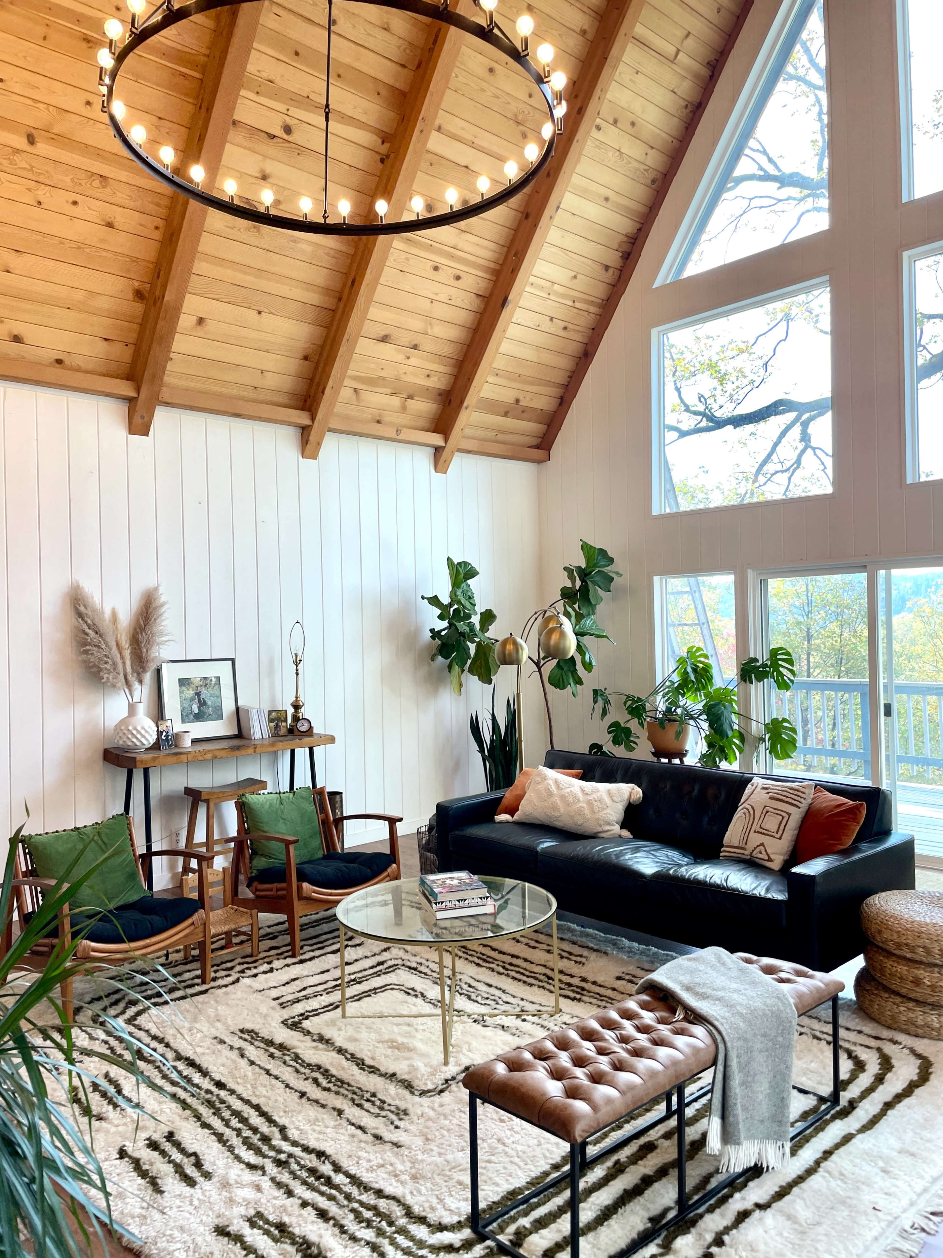The image depicts a cozy living room with a high wooden ceiling, large windows, a black leather sofa, and a glass coffee table surrounded by plants and decorative elements.