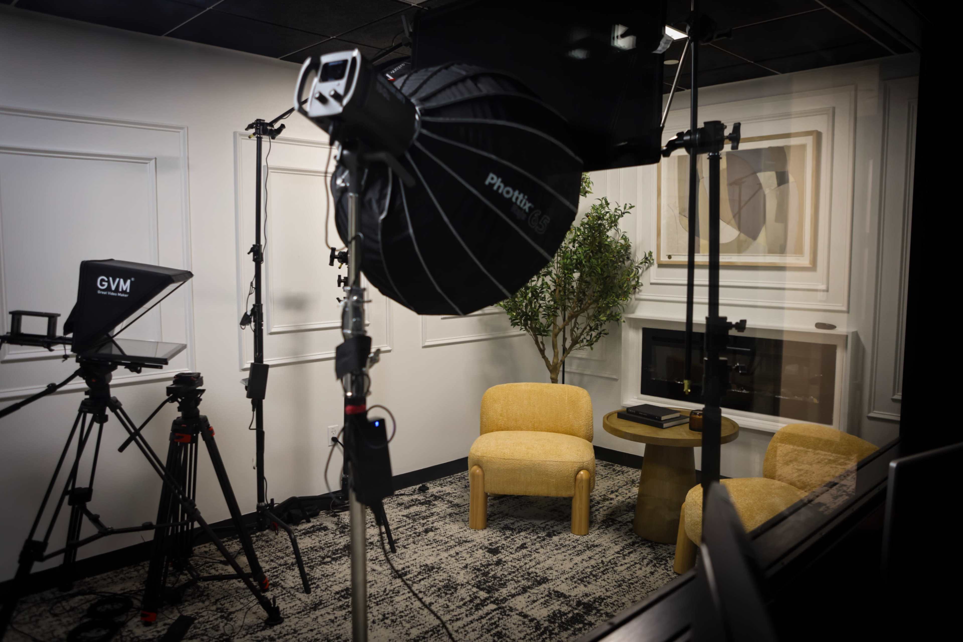 The image shows a small studio setup featuring two yellow chairs, a microphone on a stand, a camera on a tripod, and a light source, all arranged against a wall with a decorative artwork.