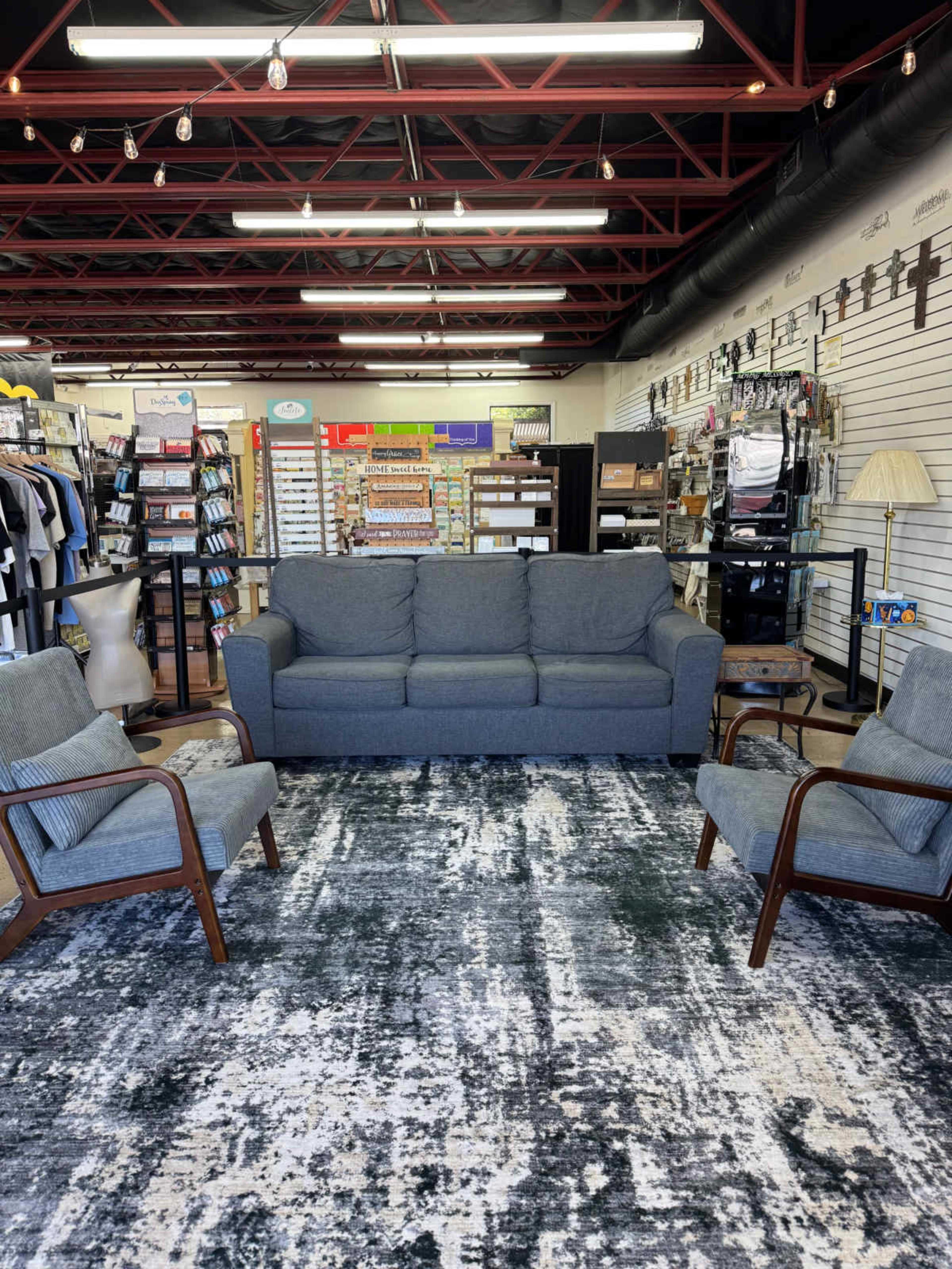 The image shows a furniture store interior featuring a gray couch and two matching armchairs arranged on a patterned rug, with various home decor items displayed on shelves in the background.