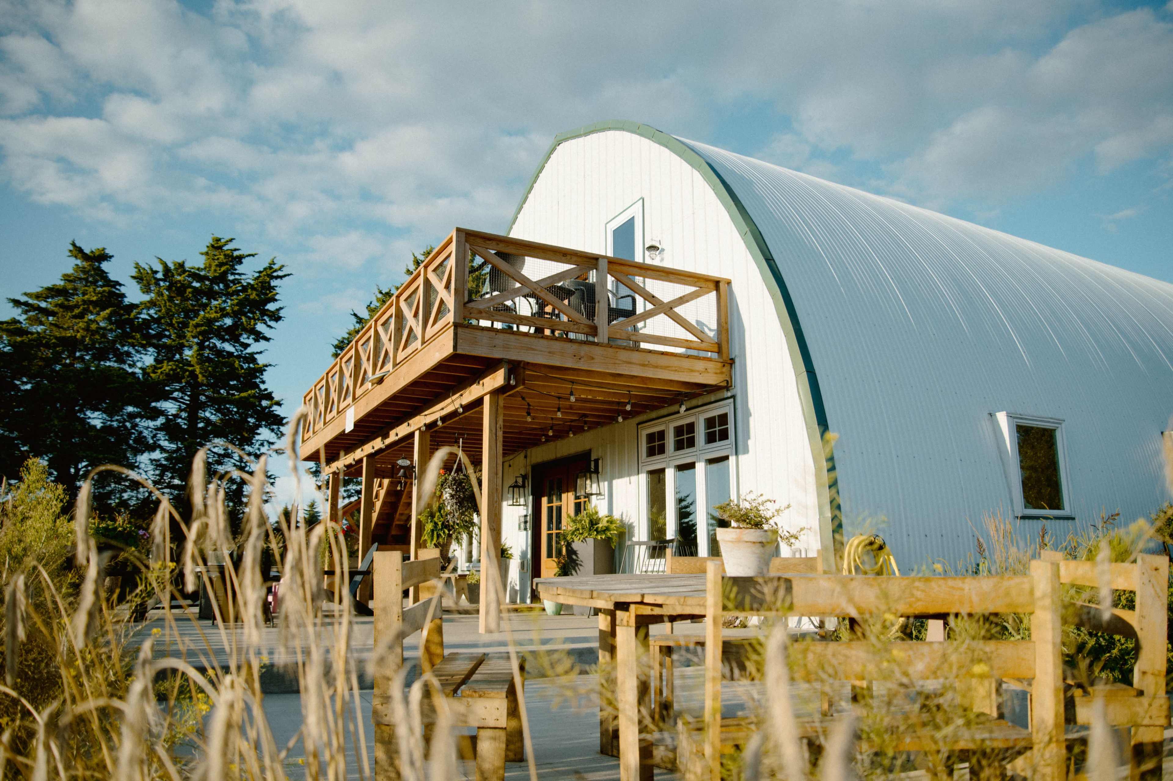 The image shows a modern, curved white building with a wooden deck and railing, surrounded by tall grass and trees under a blue sky.