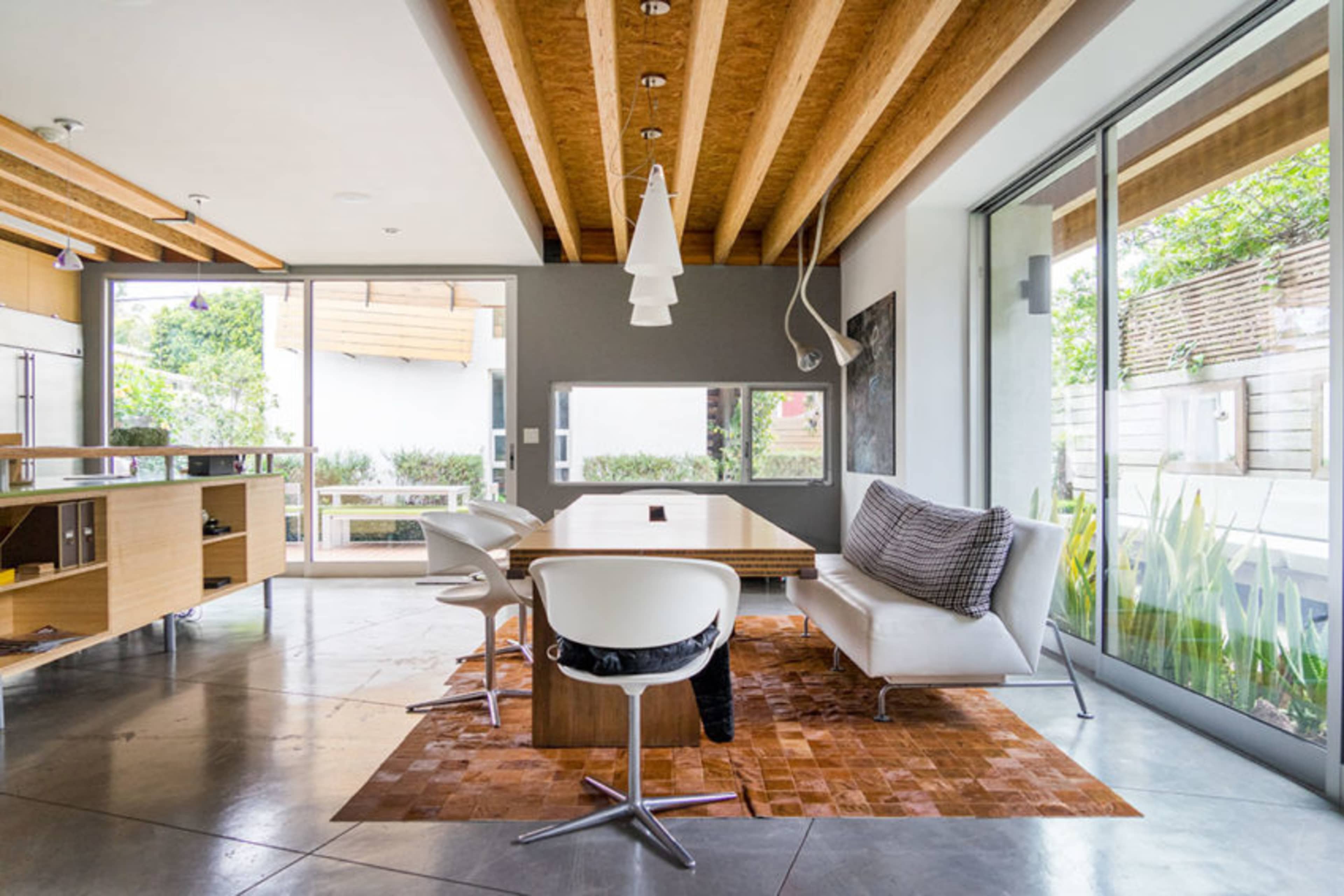 The image shows a modern dining area with a wooden table, white chairs, and large windows allowing natural light to enter.