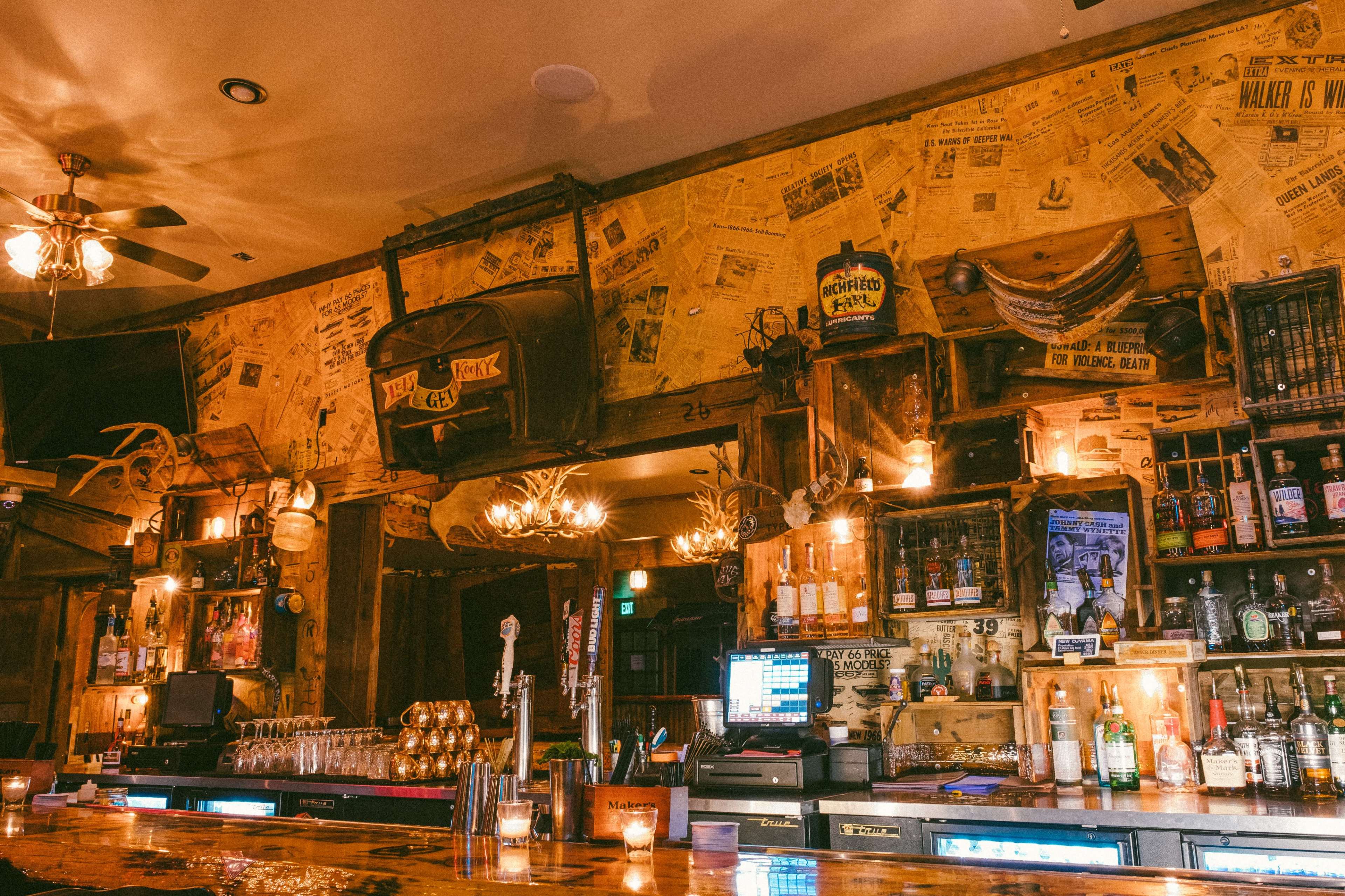 A rustic bar with a wooden countertop, shelves filled with various bottles, and walls decorated with old newspapers and other memorabilia.