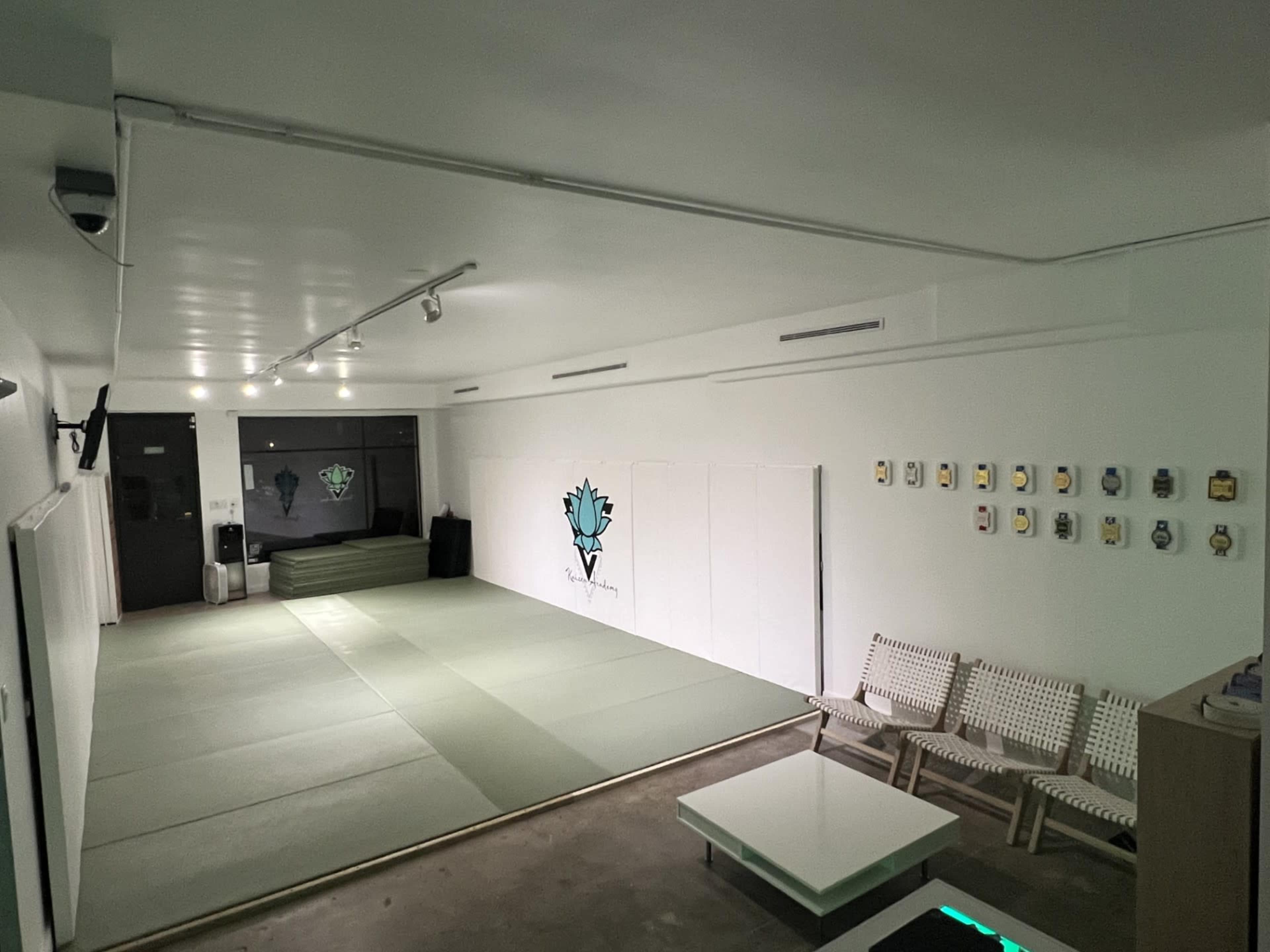 The image shows a minimalistic martial arts dojo with light green mats, a plain white wall, and a few simple chairs arranged beside a low table.