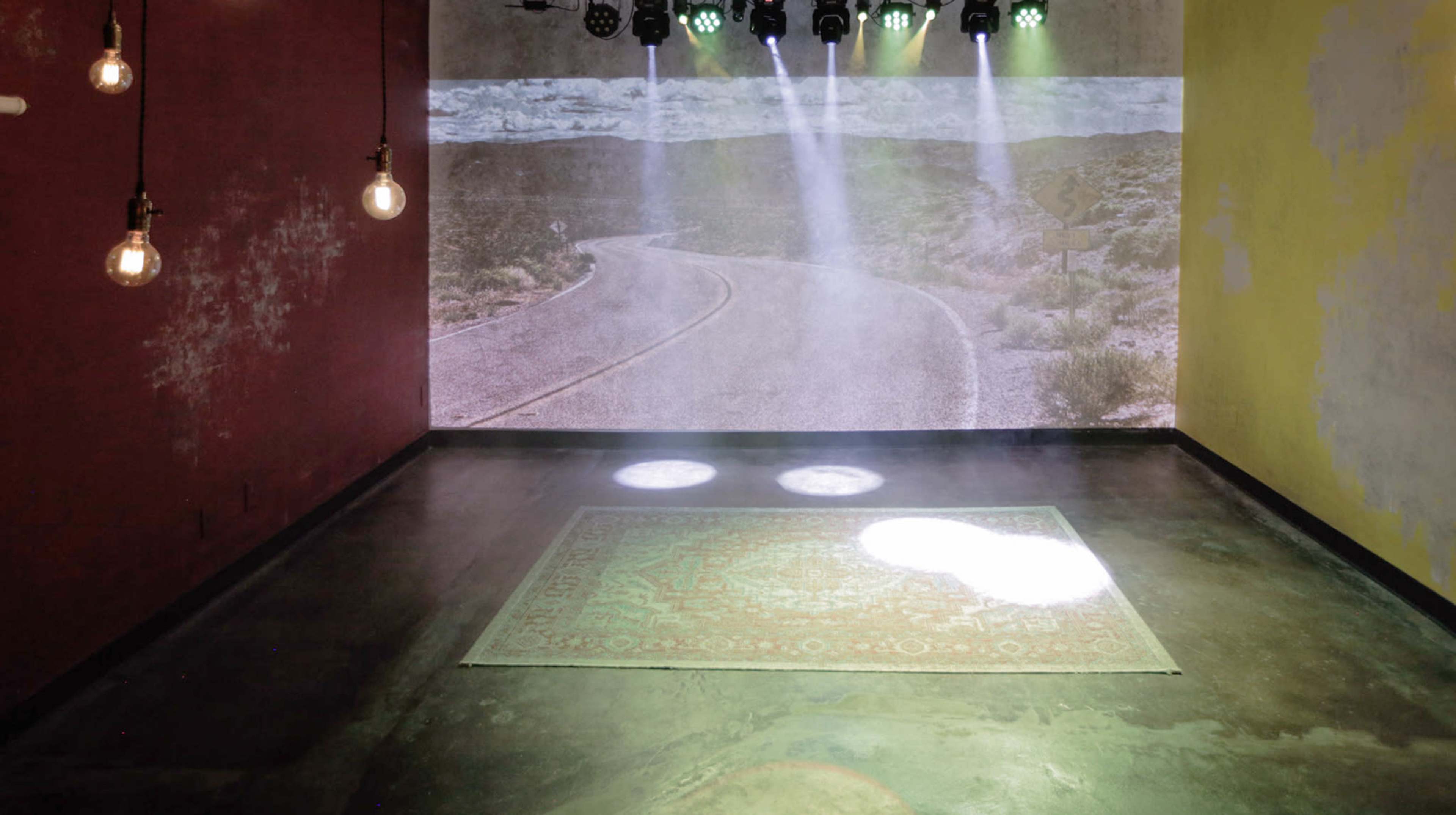 The image depicts a dimly lit room with a projected scene of a desert road on the wall and a decorative rug on the floor.