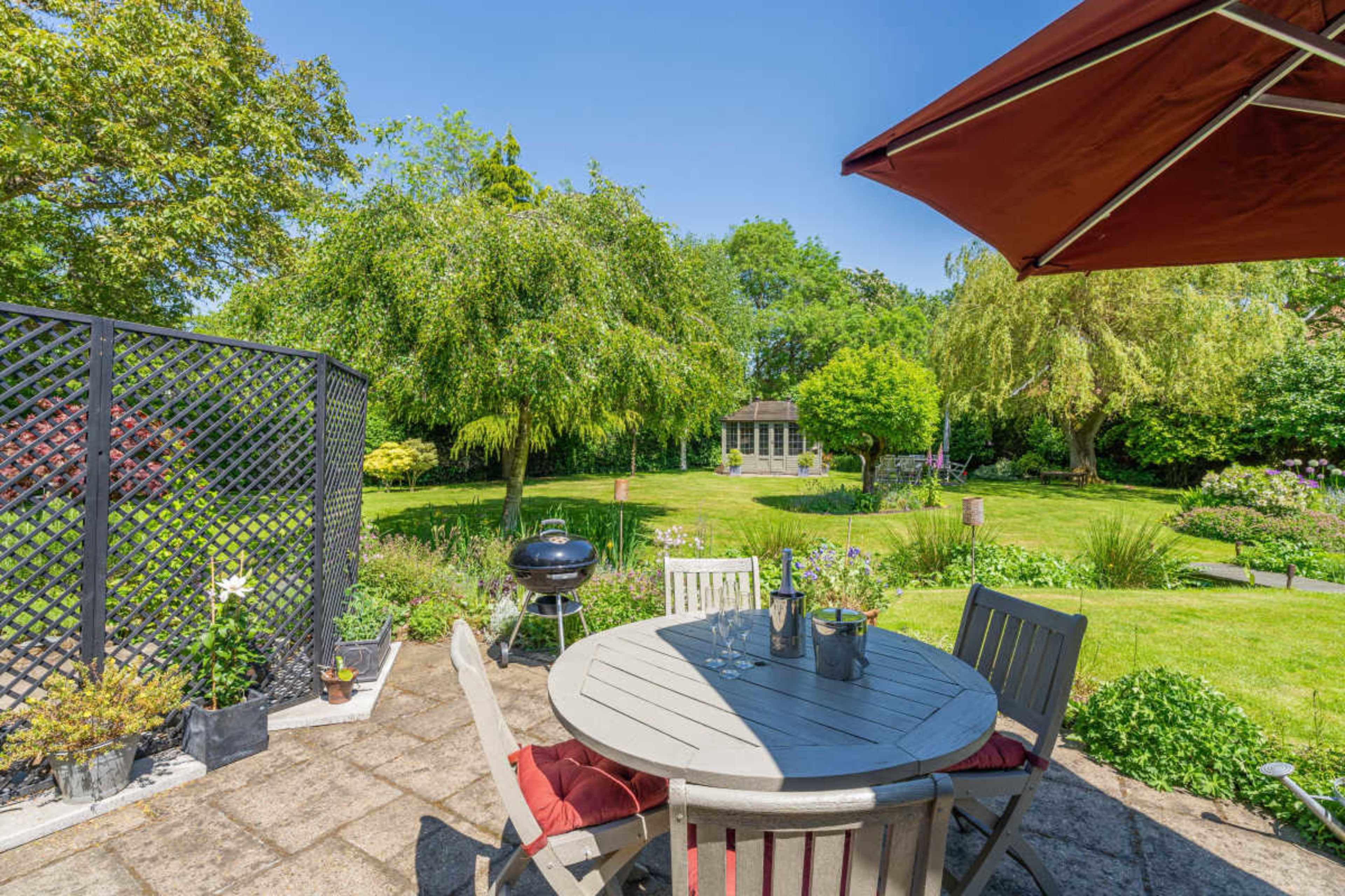 A patio area with a round table and chairs overlooks a well-maintained garden featuring various trees and greenery.