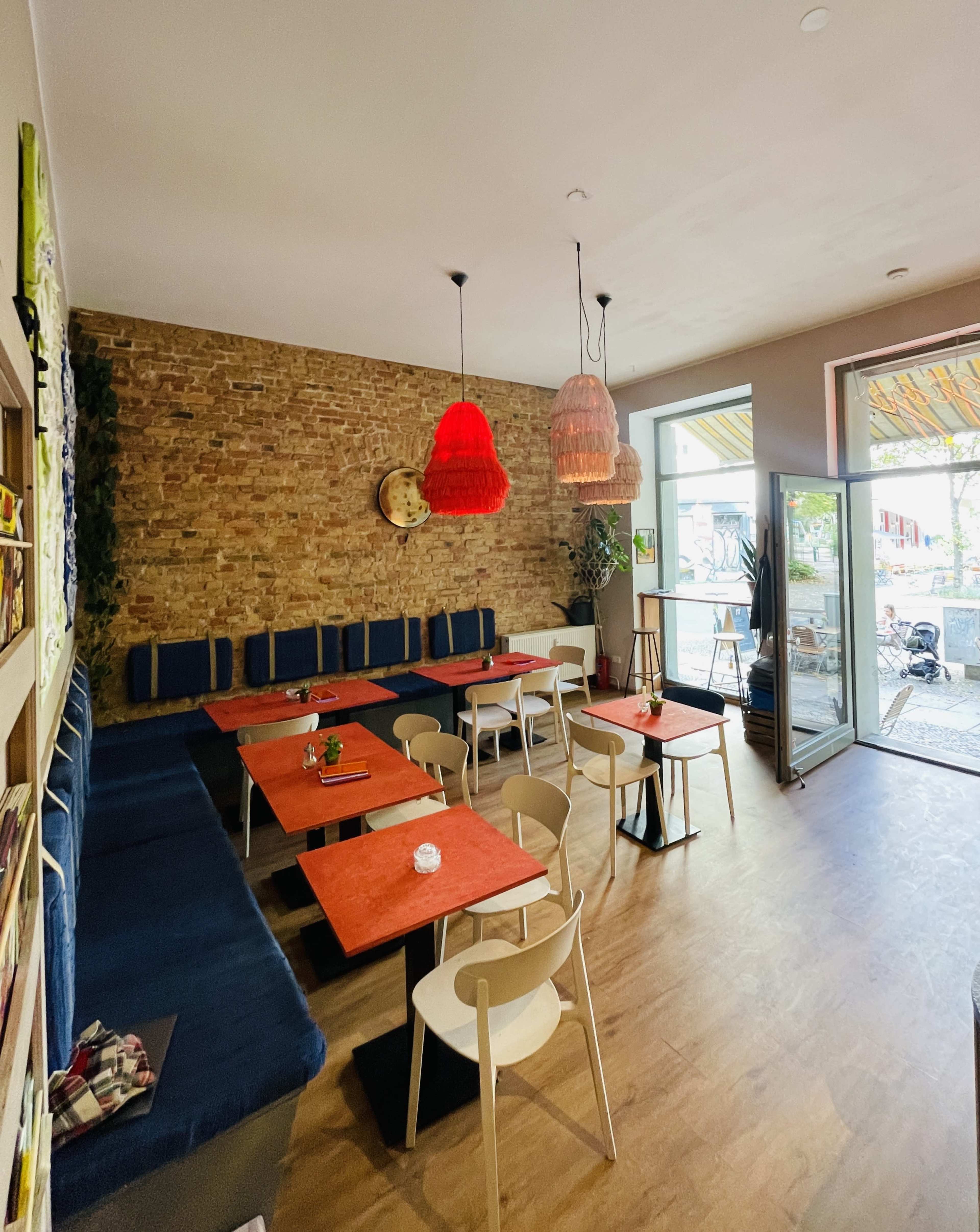 A cozy cafe features wooden flooring, brick walls, colorful pendant lights, and several tables arranged near an open entrance.