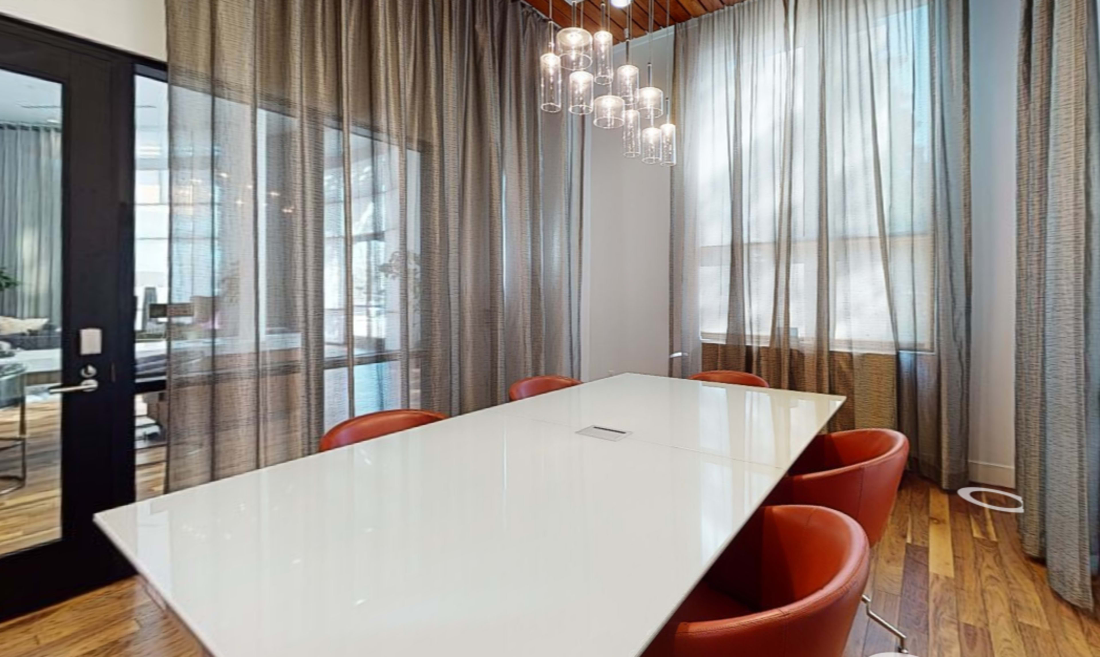 A modern conference room features a long white table surrounded by red chairs and large windows draped with sheer curtains.