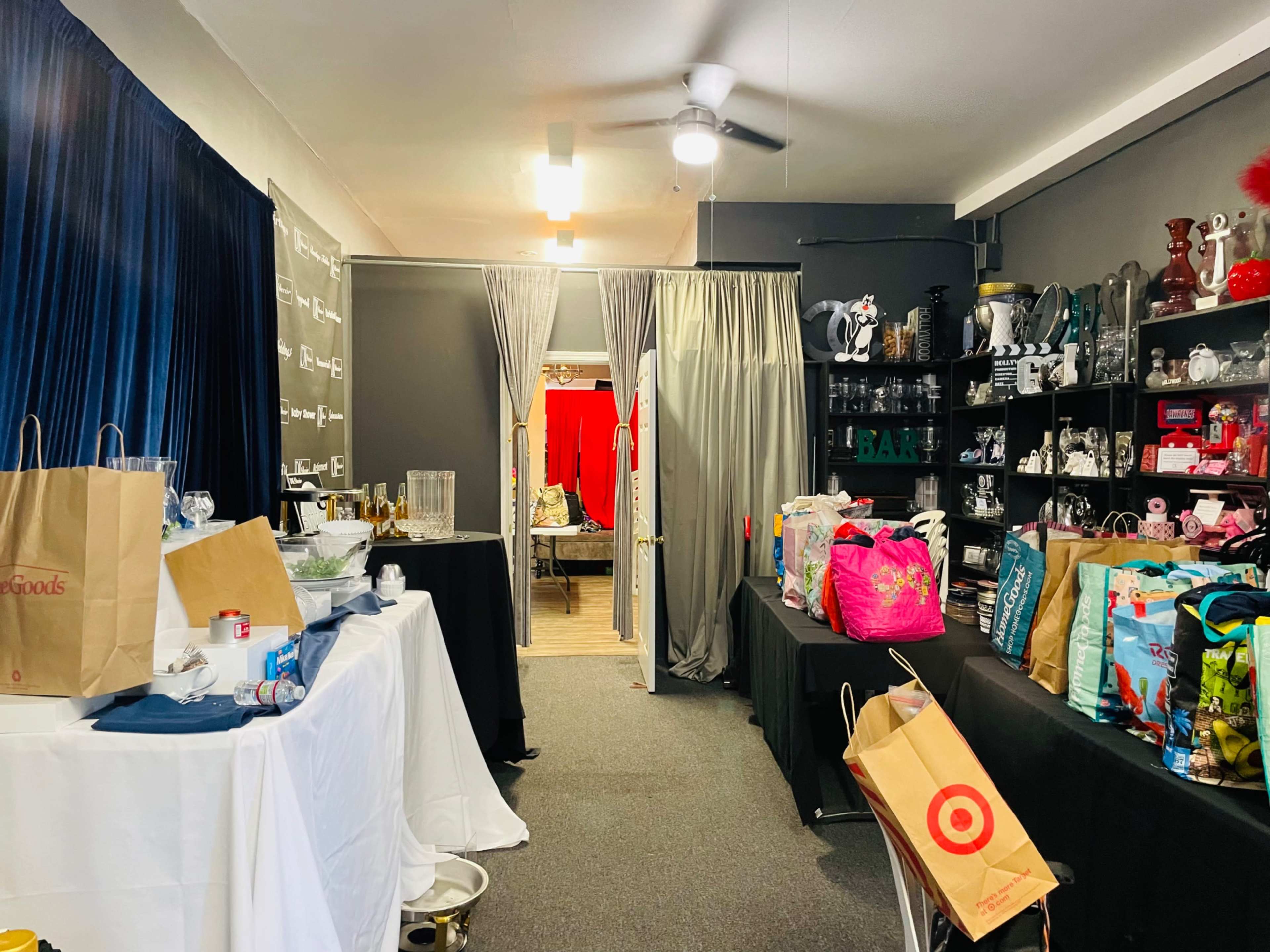 The image shows a room with shelves filled with various items, bags placed on tables, and curtains leading to another area with red decor in the background.