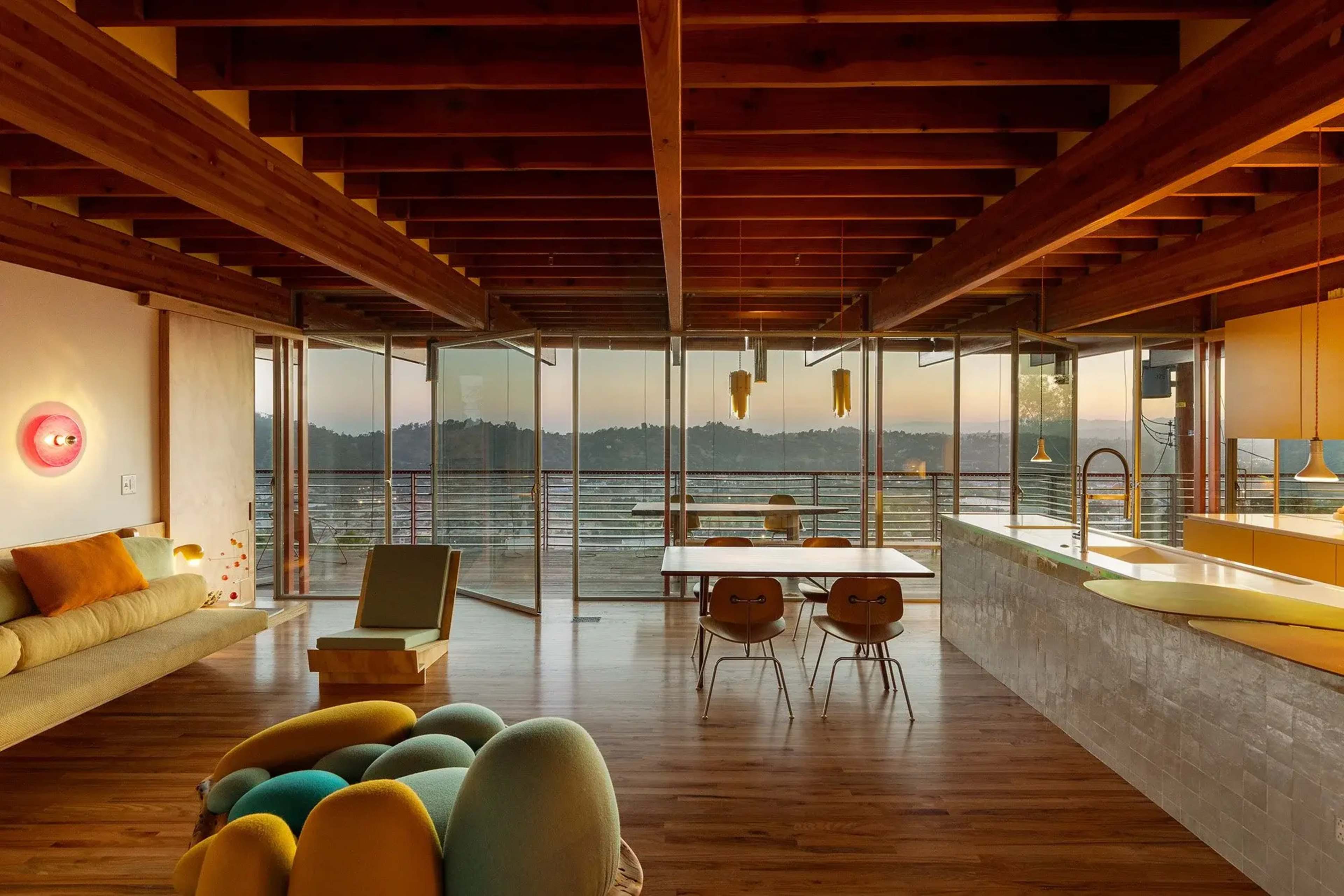 A modern living space features large glass windows, wooden beams, and a combination of cozy seating and a dining area, overlooking a scenic outdoor view.